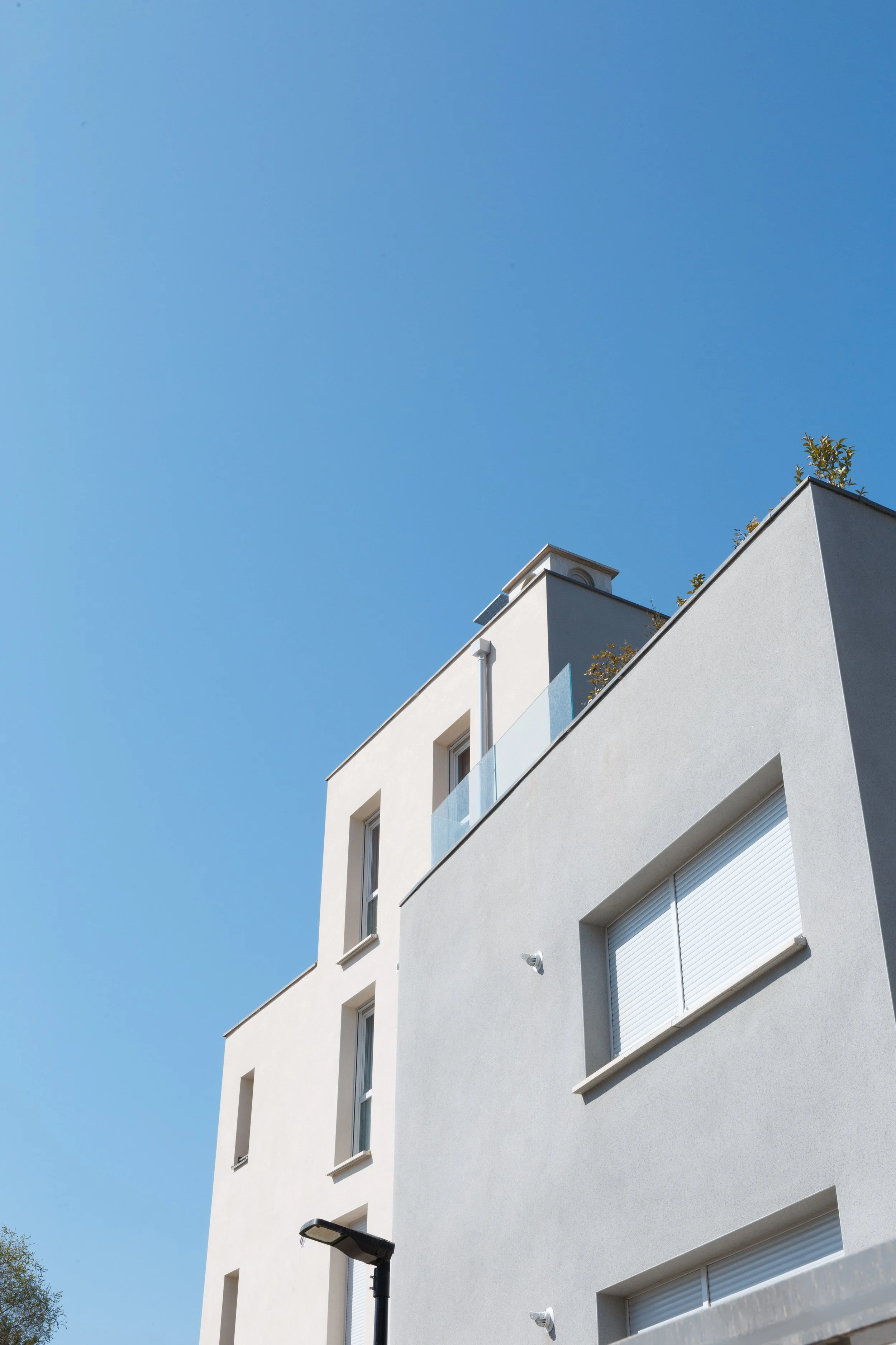 Façade d'un immeuble moderne blanc avec plusieurs fenêtres à volets roulants fermés, un lampadaire street style et un ciel bleu clair.