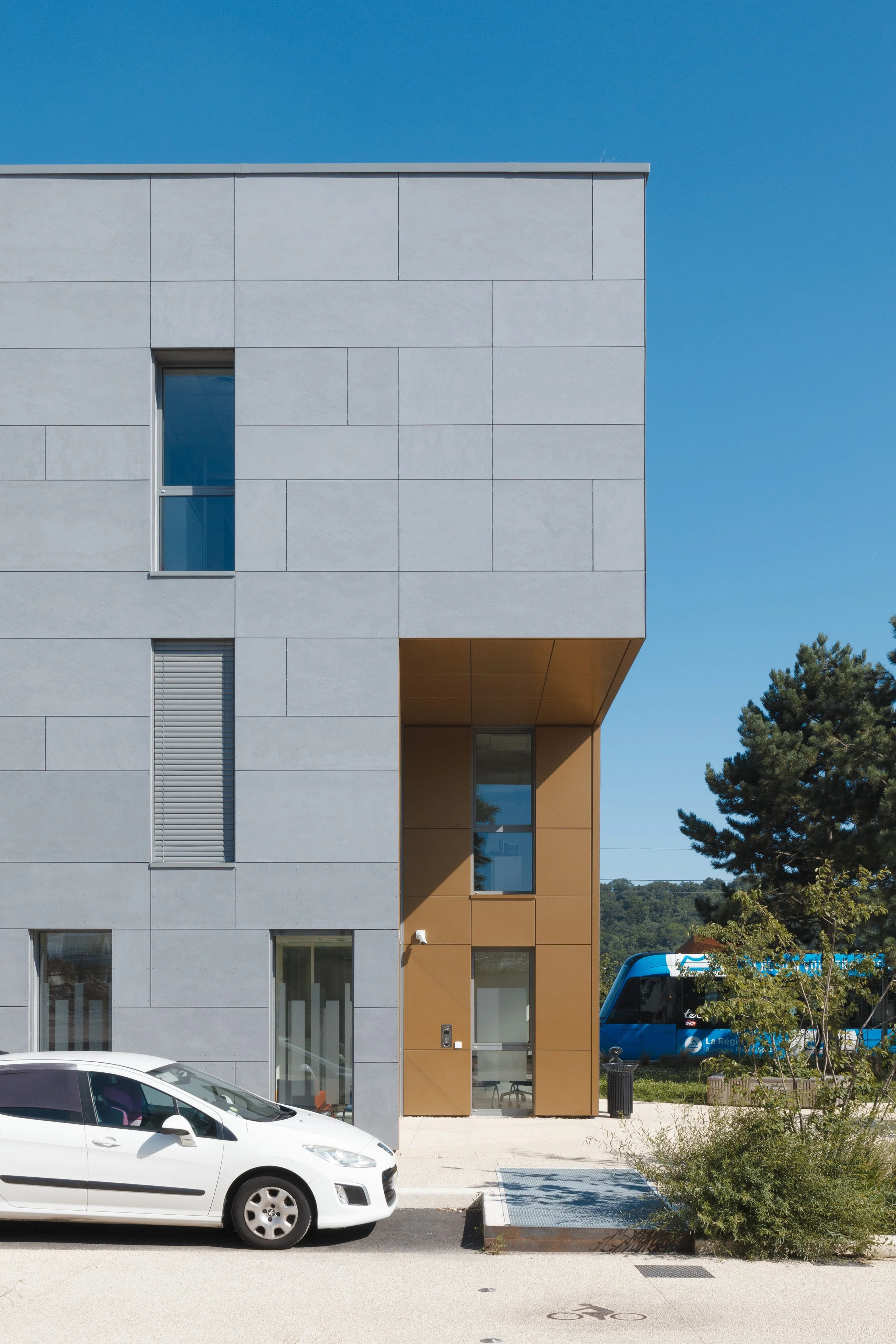 Bâtiment moderne avec façade en pierre claire, fenêtres rectangulaires et un ciel bleu clair en arrière-plan. Une voiture blanche est garée devant le bâtiment, avec une zone pour vélos en face.