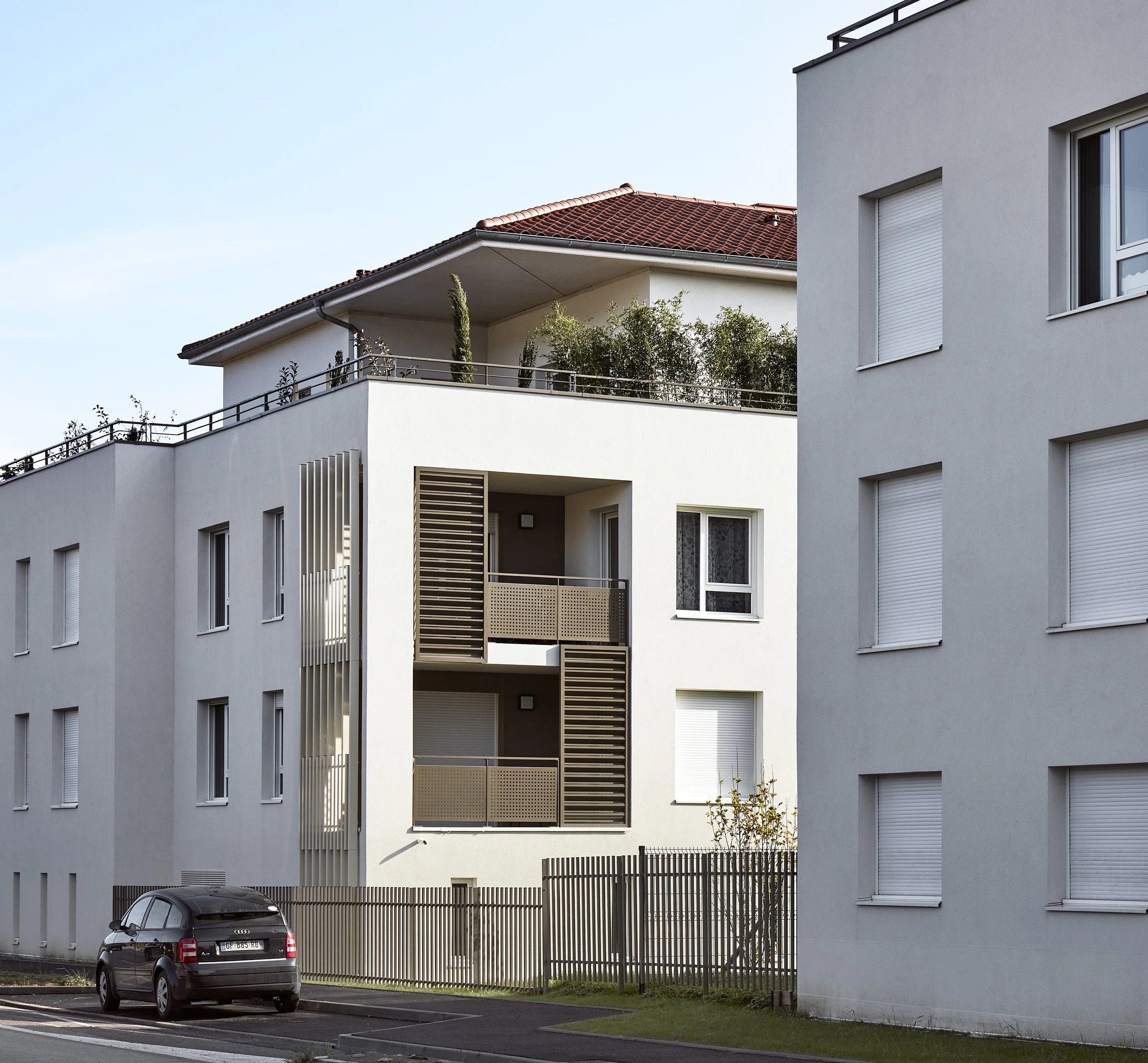 Appartement moderne avec plusieurs fenêtres à volets roulants blancs, balcon avec panneau de pare-vue en métal perforé, et parking avec voiture noire en première plan.