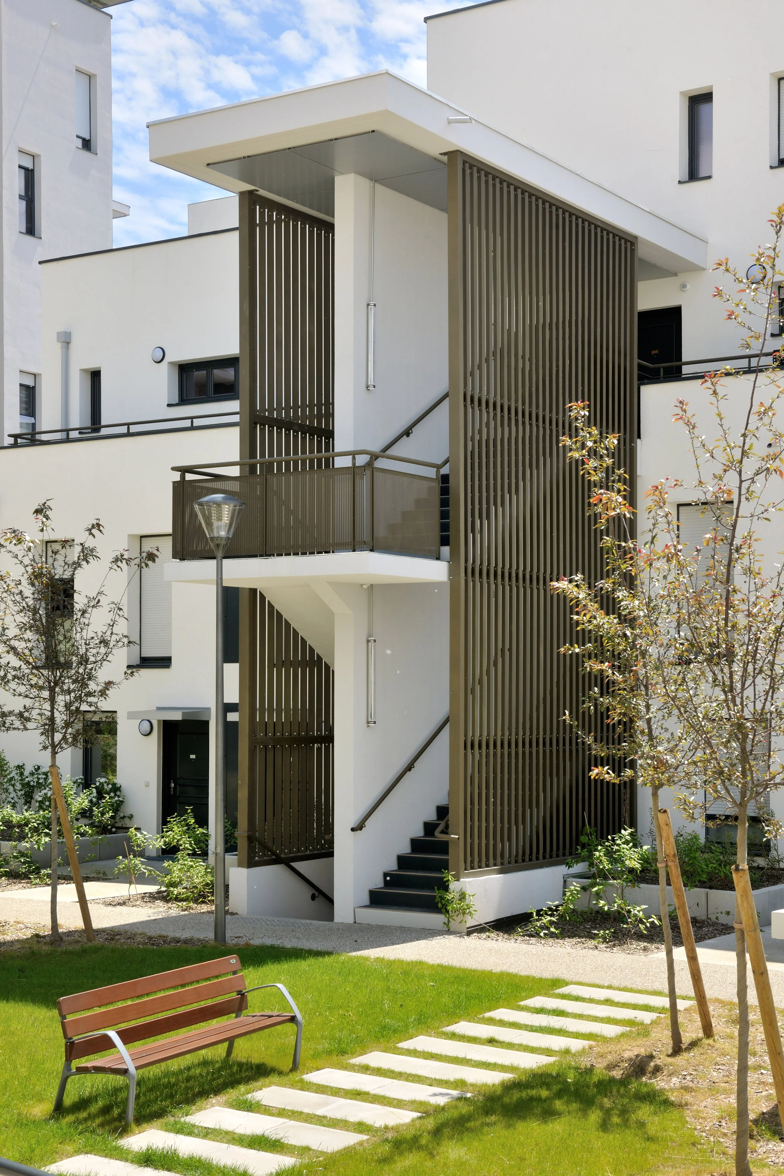 Appartement moderne blanc avec balcon en aluminium noir, escaliers extérieurs et jardin avec arbres et pelouse.