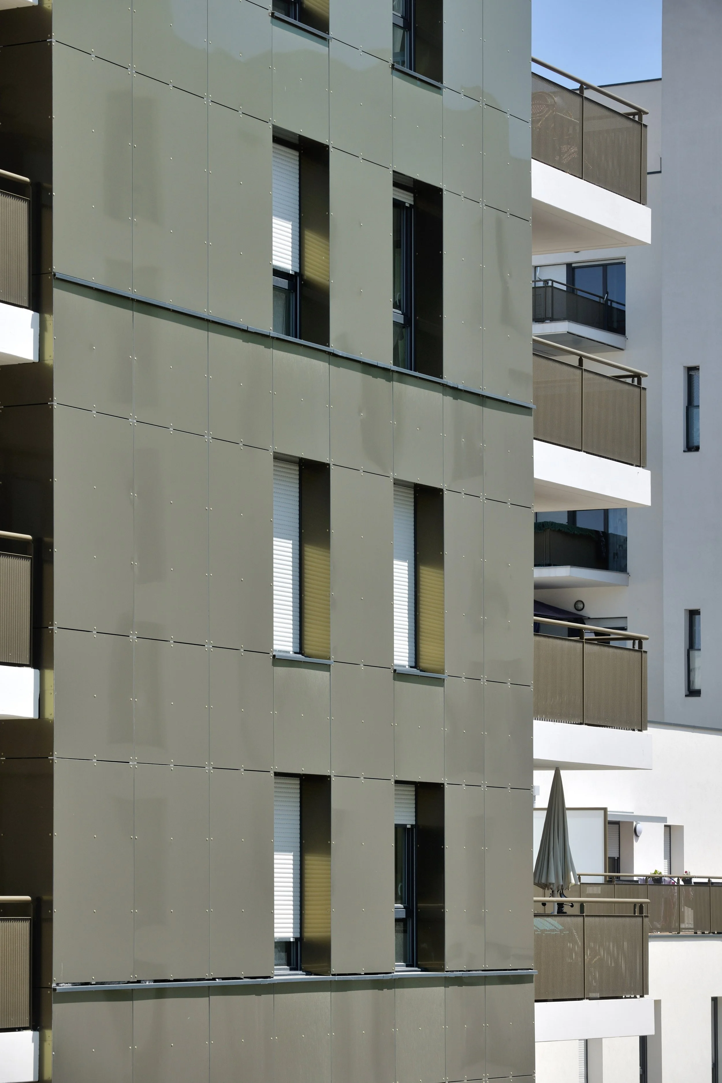 Façade moderne d'un immeuble avec des balcons et des fenêtres à volets roulants, avec revêtement en panneaux vert grisâtre.