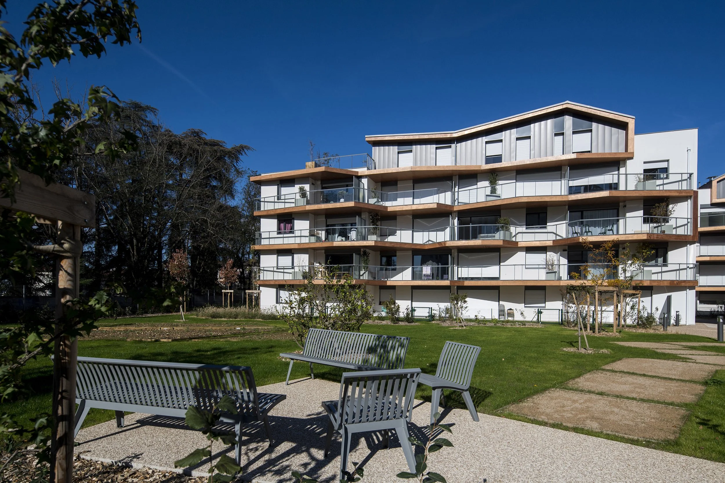 Appartement moderne avec balcons en étage supérieur, dans un environnement verdoyant avec des bancs dans un parc, ciel clair