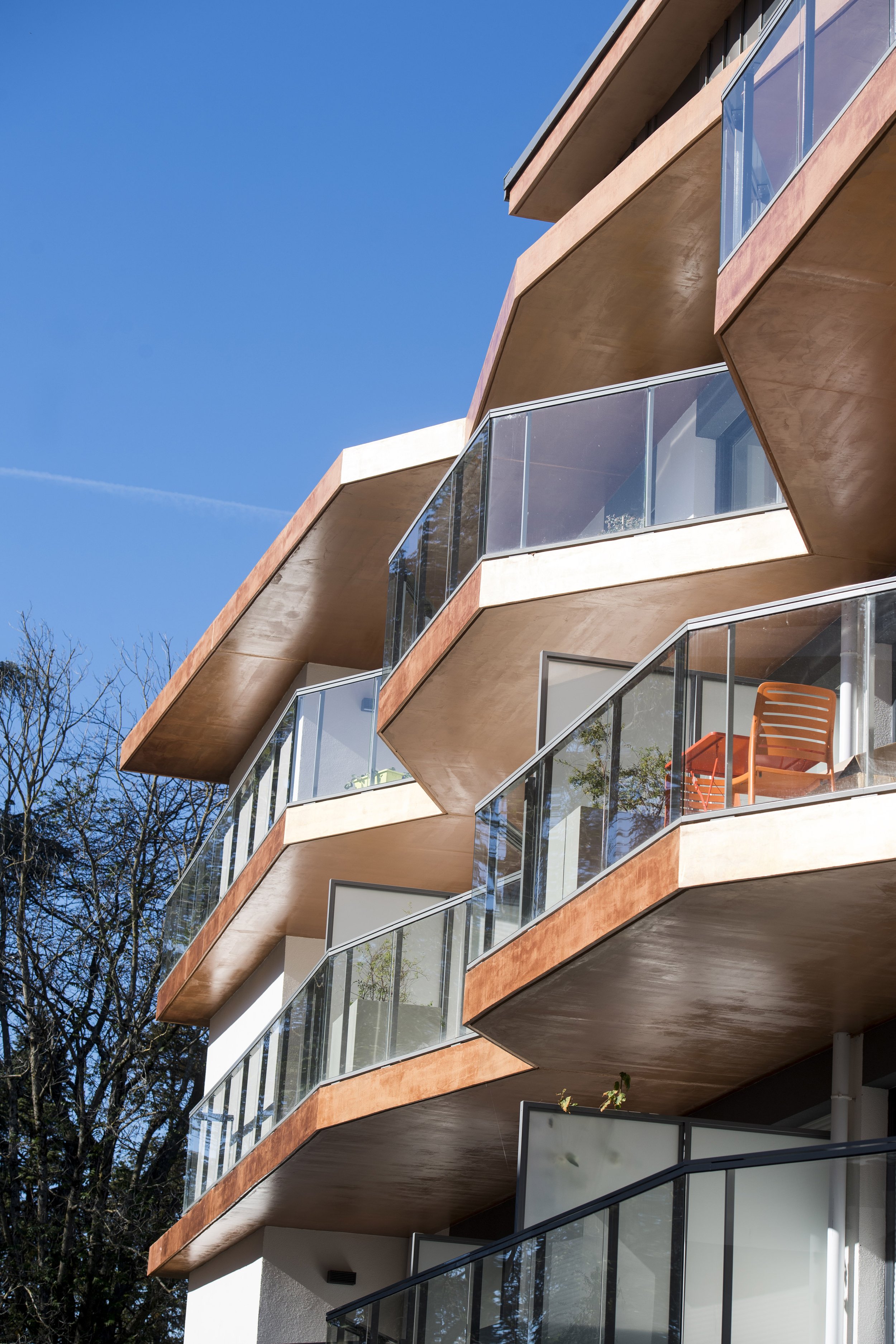 Balcon d'un bâtiment résidentiel moderne avec des parois en verre, façades en béton ardoise, et un ciel bleu en arrière-plan.