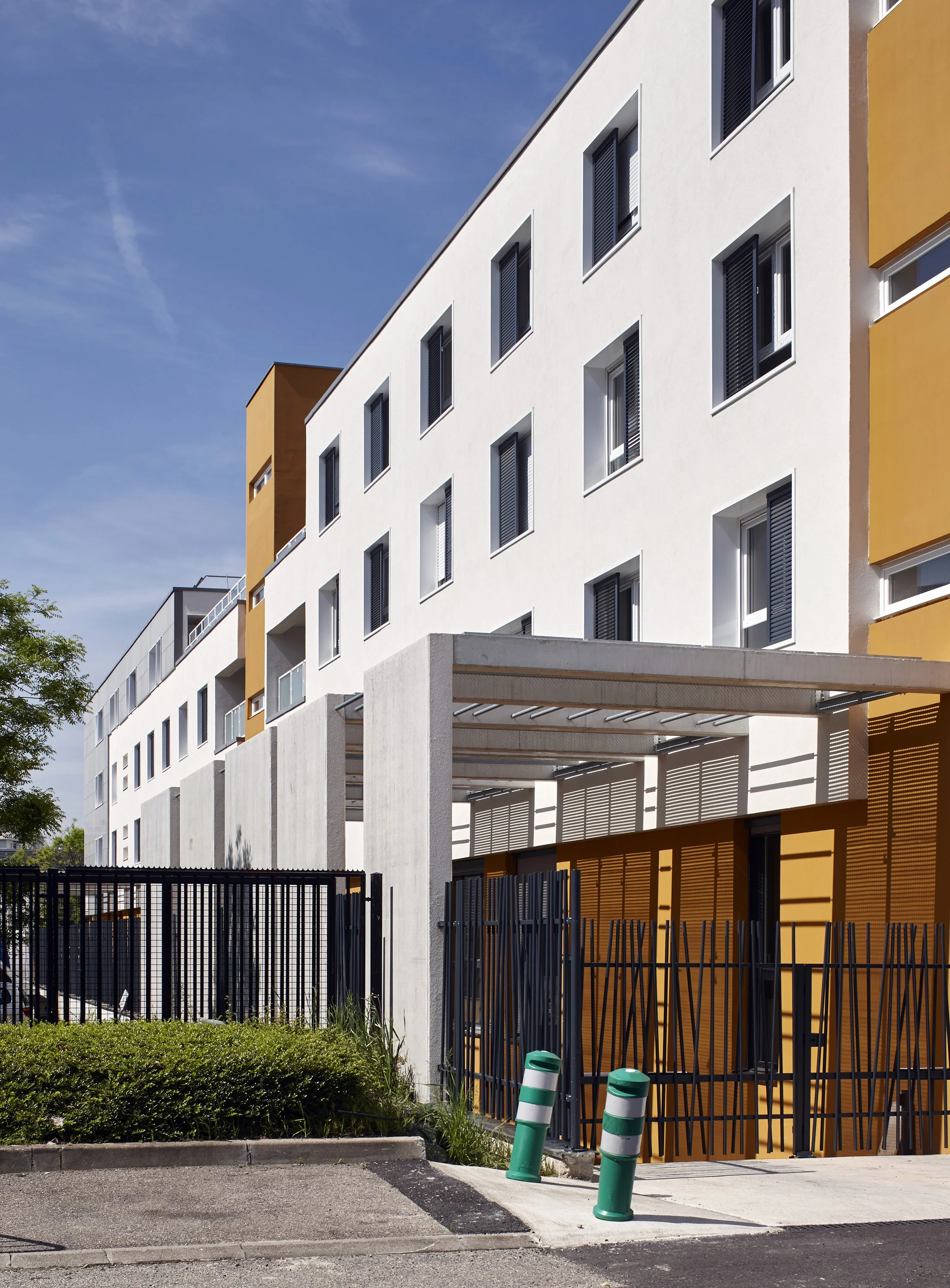 Résidence moderne avec façade blanche et accents en jaune, portes couvertes d'ombrages, clôture en métal noire, et espaces verts avec quelques piétons, sous un ciel bleu avec quelques nuages.