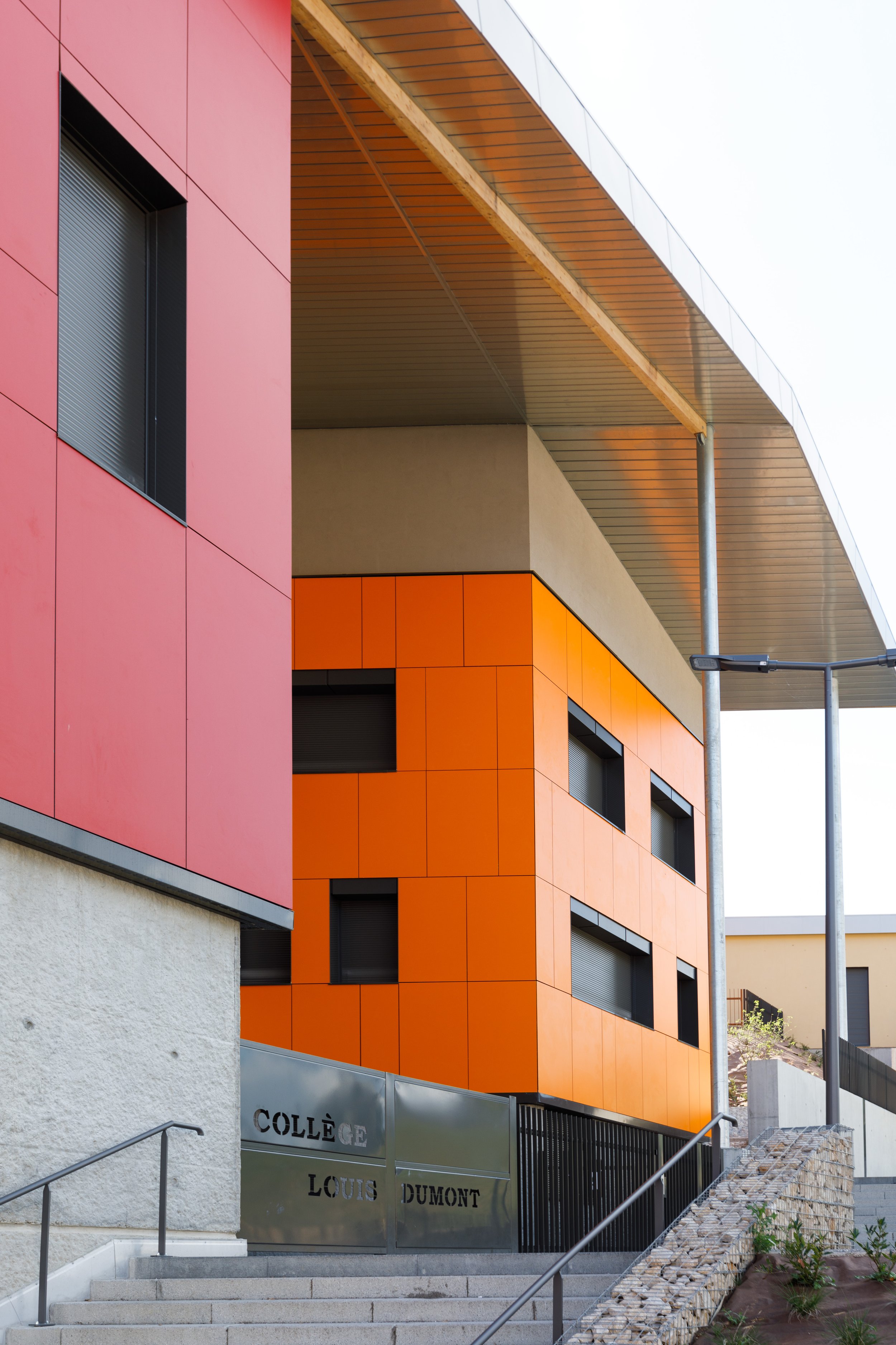 Façade moderne colorée d'un bâtiment avec panneaux orange et rose, entrée avec escaliers et panneau en métal portant l'inscription 'Collège Louis Dumont'.