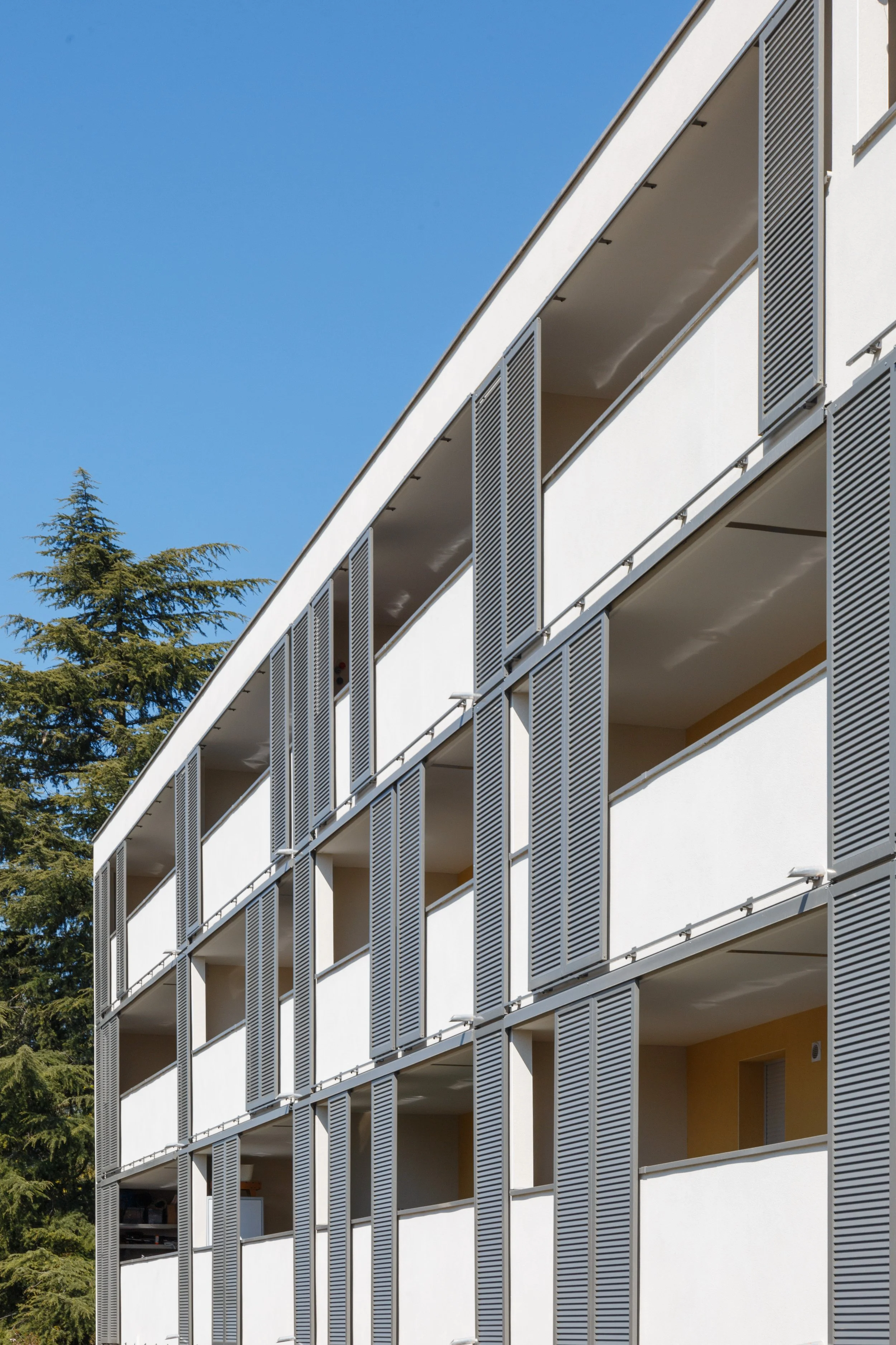 Vue d'un bâtiment moderne avec plusieurs balcons, volets en lamelles métalliques gris, et un ciel bleu clair en arrière-plan. Il y a aussi un arbre à gauche.