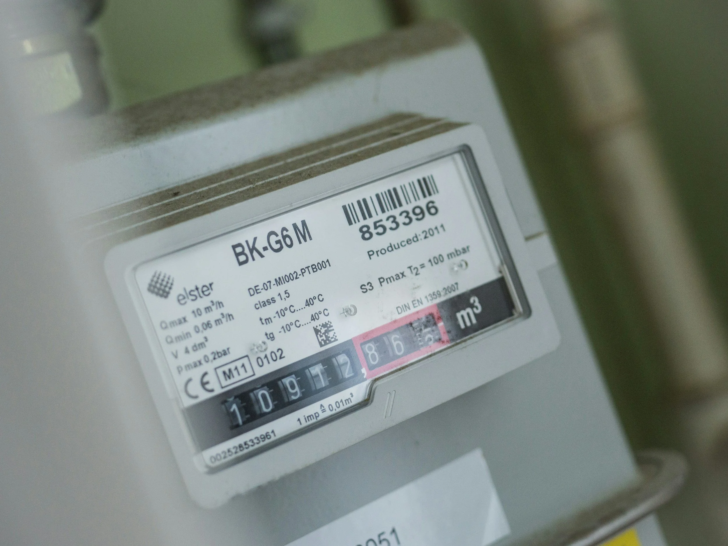 A close-up of a digital gas meter with a white display panel showing various readings and information, mounted on a wall with a green pipe visible at the top.