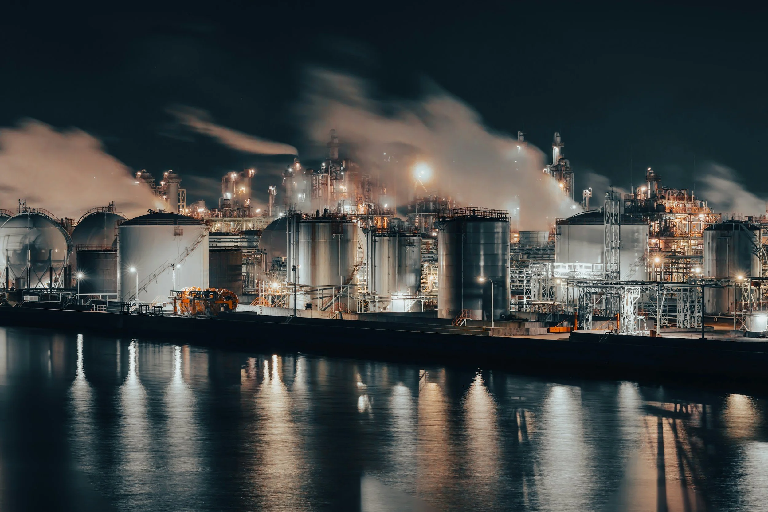 Nighttime industrial refinery with large cylindrical tanks, smoke emissions, and bright lights reflecting on water in the foreground.