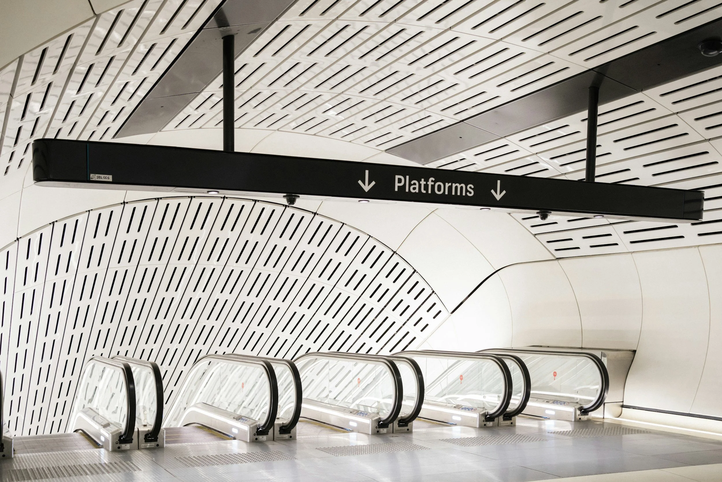 Empty escalators in a modern underground subway station with white and black curved ceiling panels and a black sign overhead with white text reading "Platforms" and downward arrows.