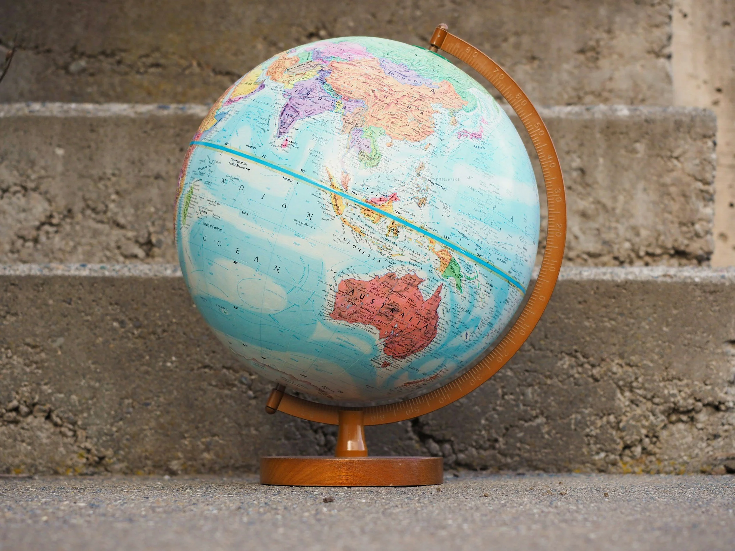 A globe showing continents such as Asia, Australia, and parts of Africa, with an orange semi-meridian at the side, resting on a brown base on a textured concrete surface.