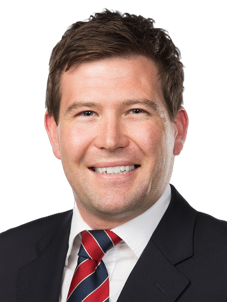 A man with short brown hair, wearing a dark suit, white shirt, and a red striped tie, smiling against a plain background.