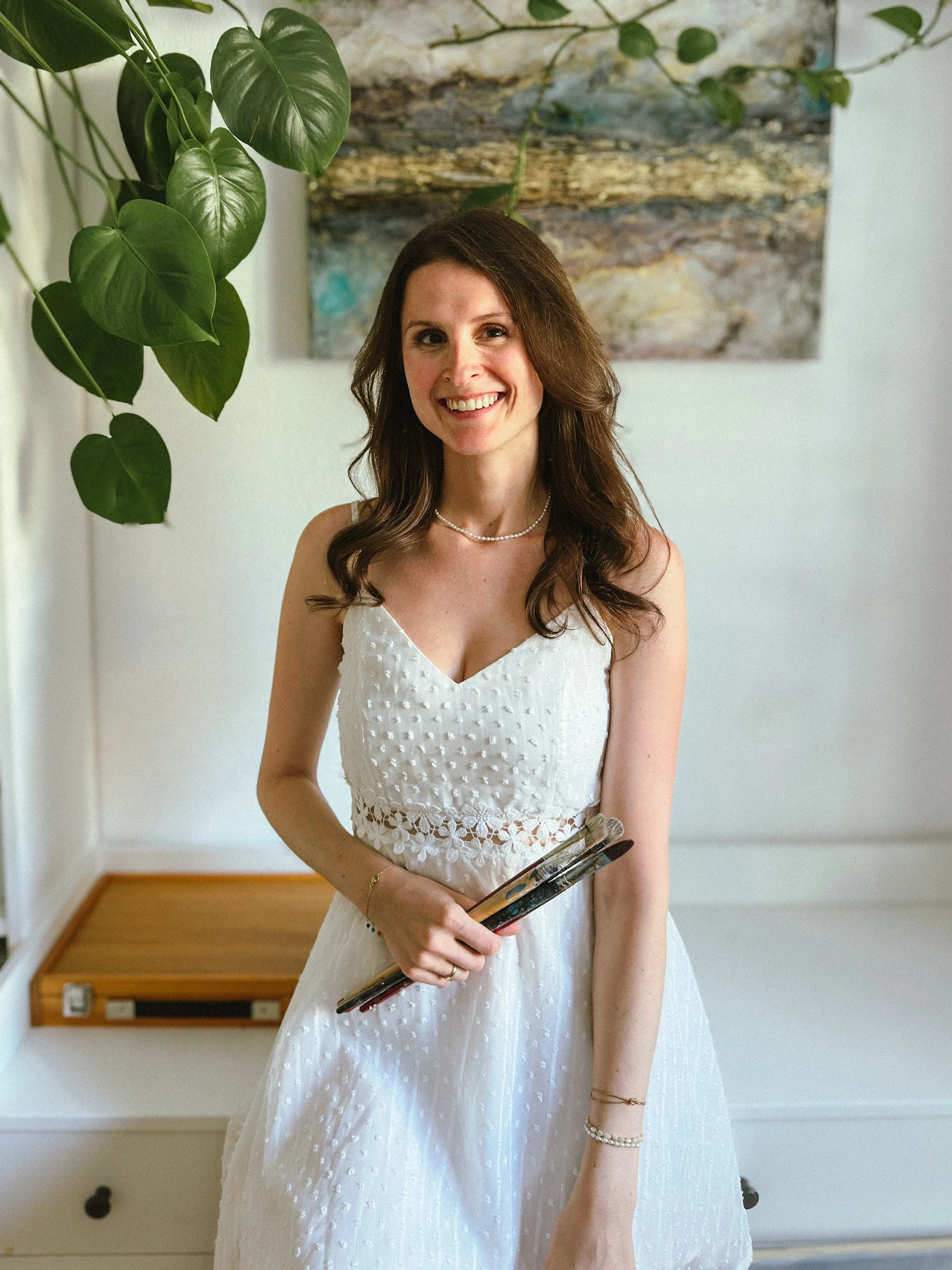 A woman in a white dress holding glasses, standing indoors near a green plant and a painting on the wall while smiling.