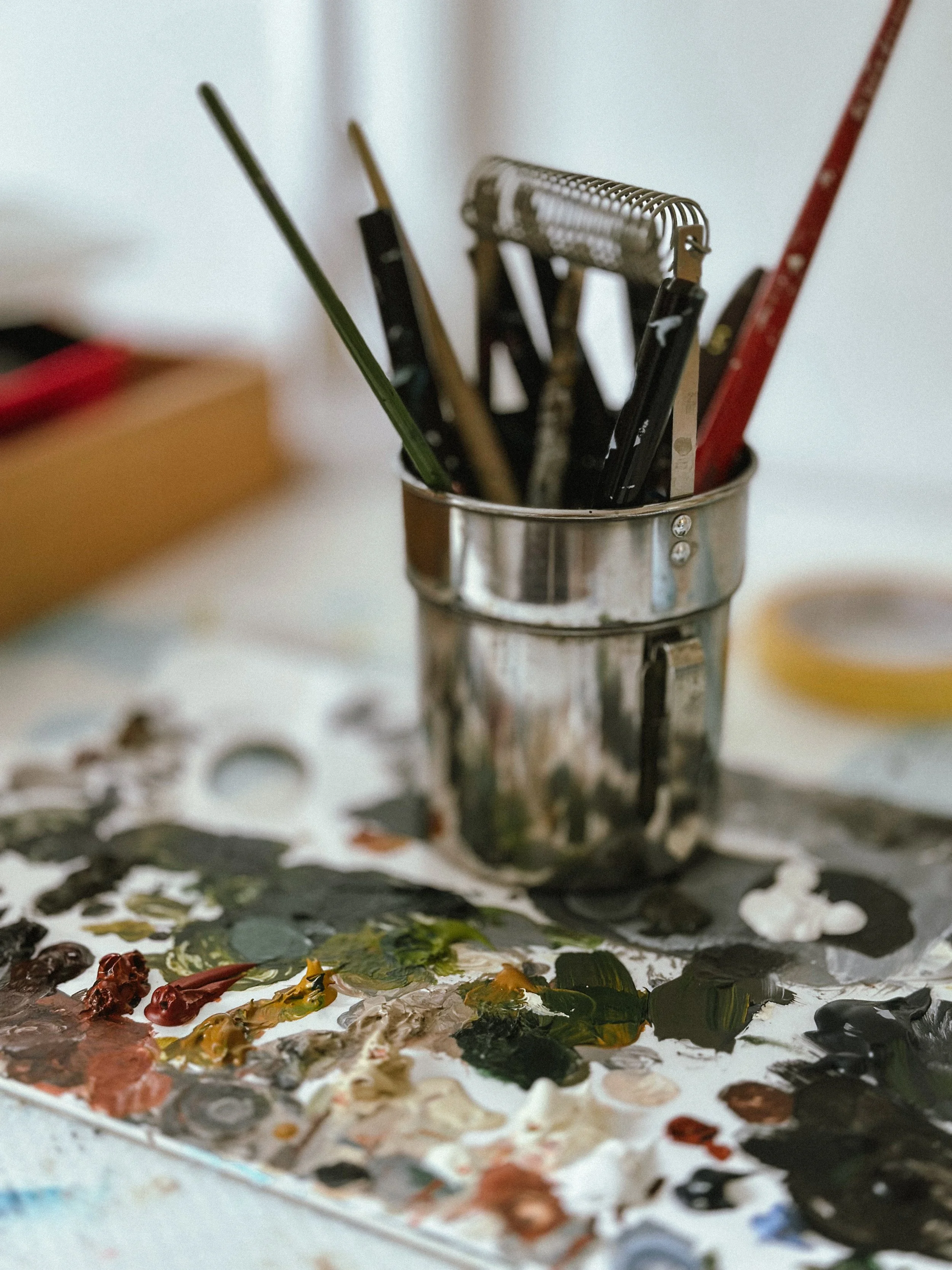 A paint palette with mixed colors and a container holding various paintbrushes and tools used for painting.