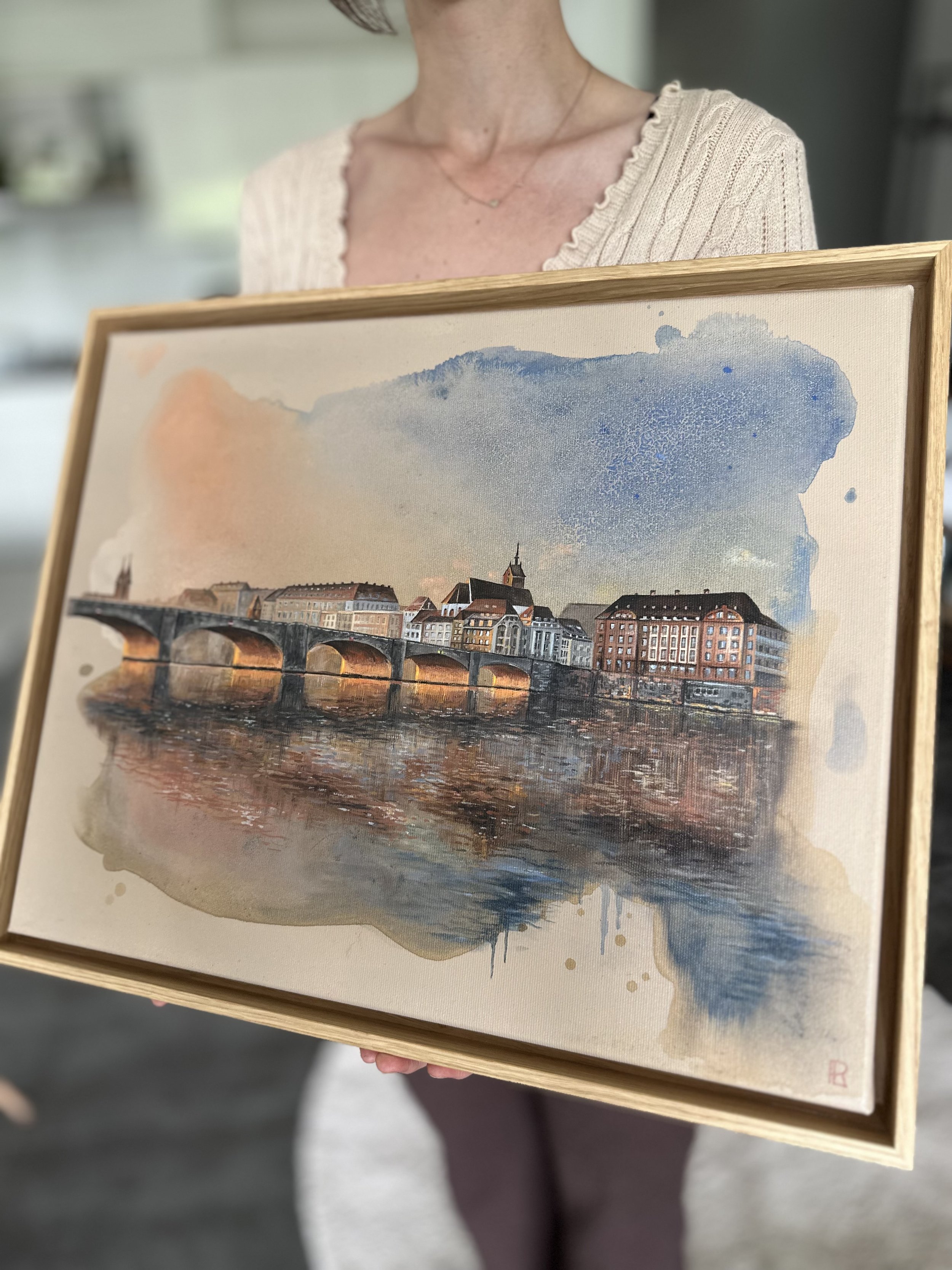 Person holding framed watercolor painting of a European cityscape with bridge and buildings by a river.