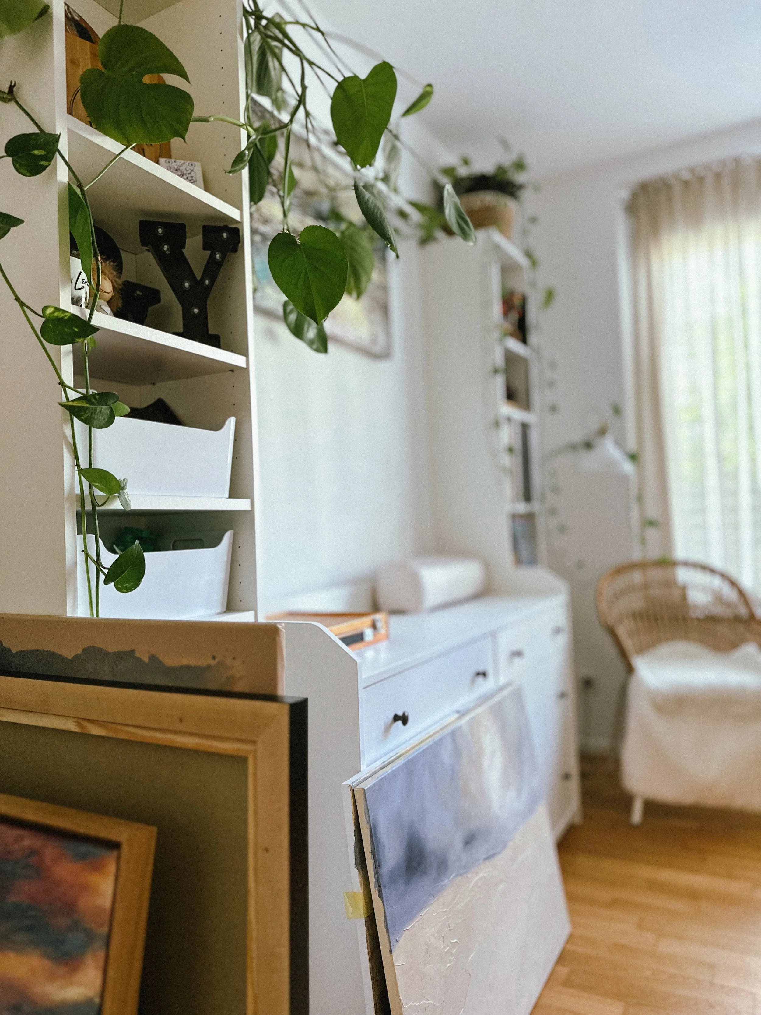 A room with a white desk holding an unfinished painting of a landscape, a white wicker chair nearby, and shelves with various plants and decorative items, natural light coming through a large window with curtains.