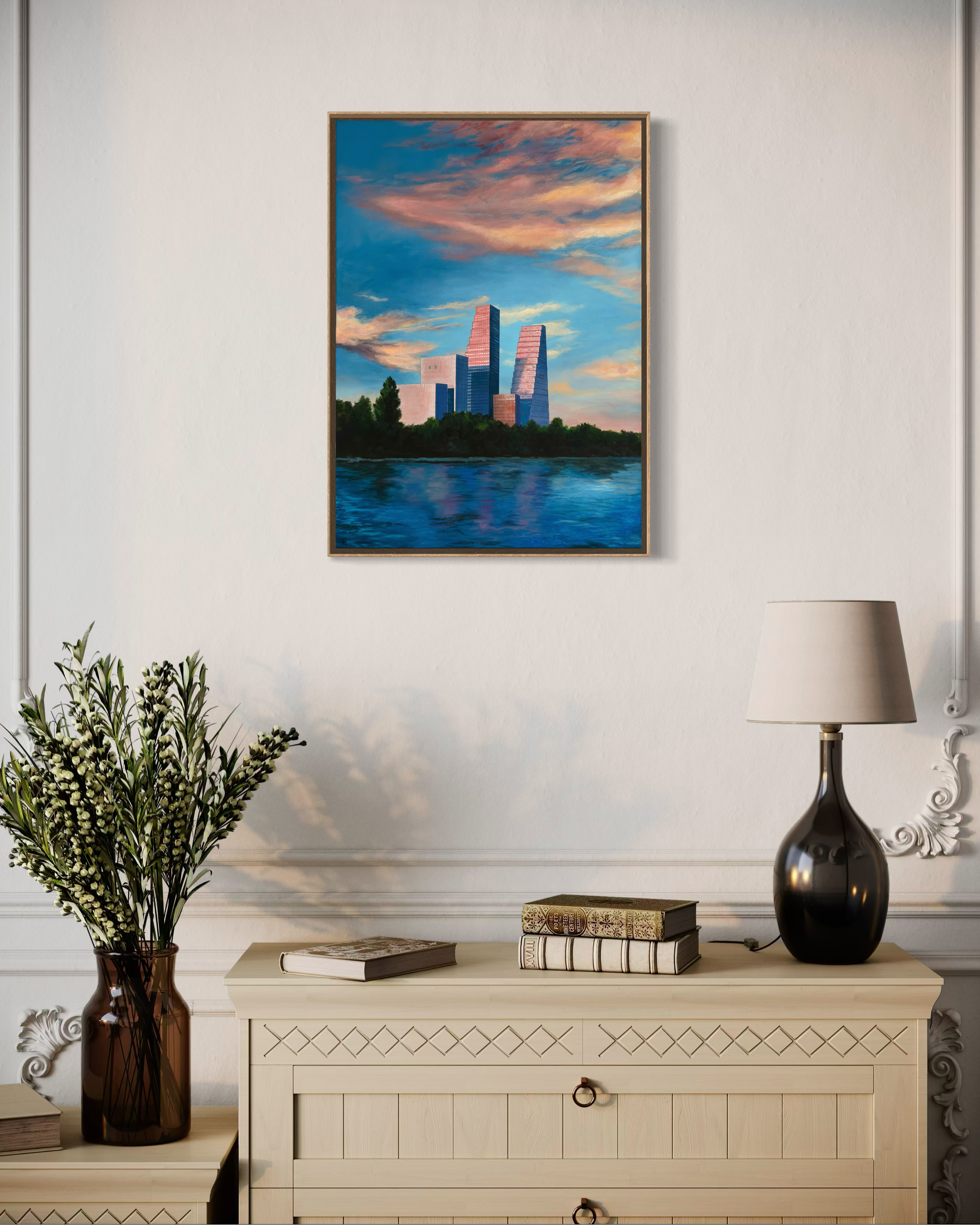 A framed painting of a sunset over buildings and water hangs on a white wall. Below it, there is a beige sideboard with decorative handles. On top of the sideboard are three books and a black and gold decorative lamp. To the left of the sideboard, there is a tall vase with green and white flowering plants. The wall has ornate white molding, and part of a white table lamp and a small decorative sign are visible.