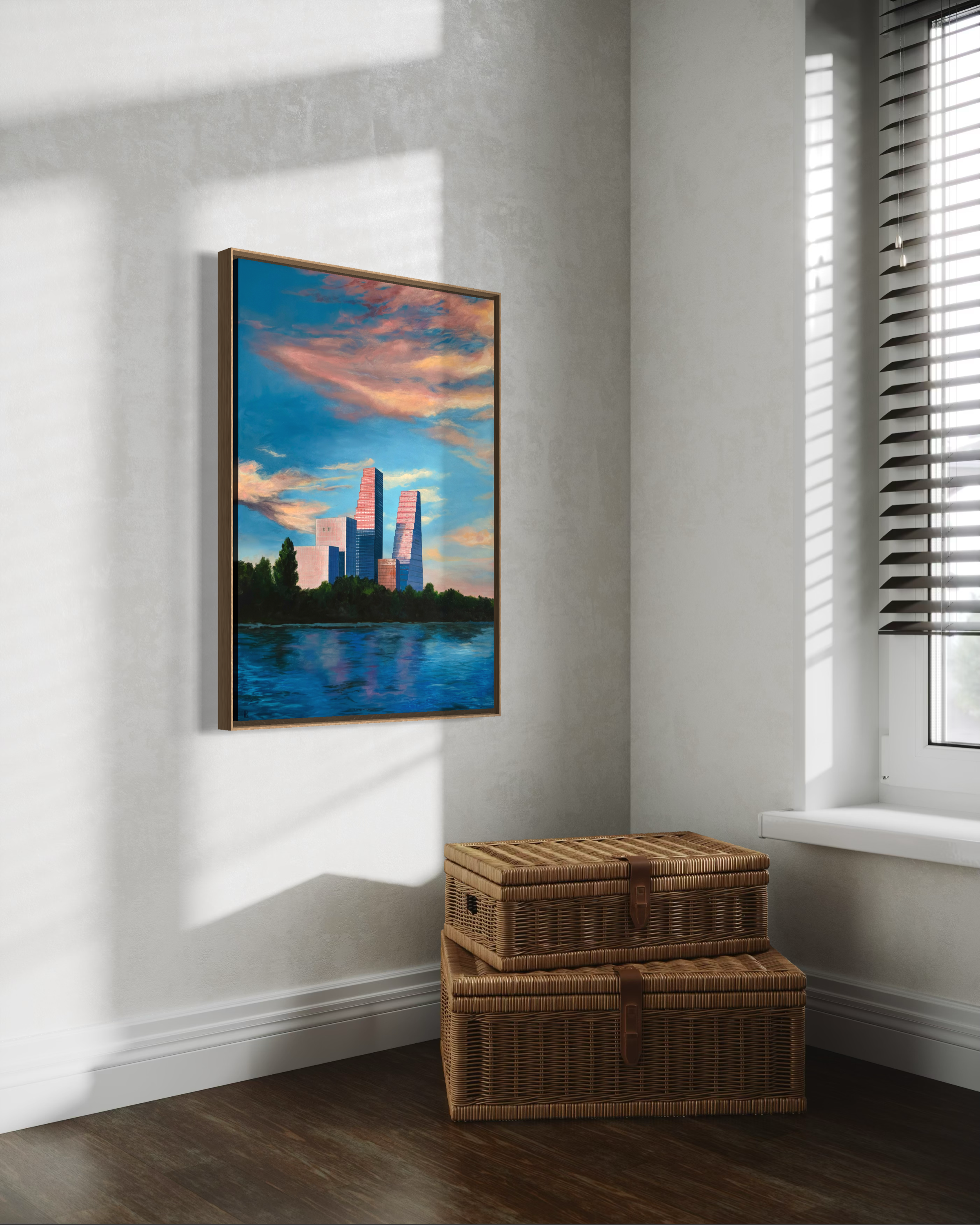 A corner of a room with a wicker storage chest, a framed landscape painting, and a window with horizontal blinds casting shadows on the white wall and dark hardwood floor.