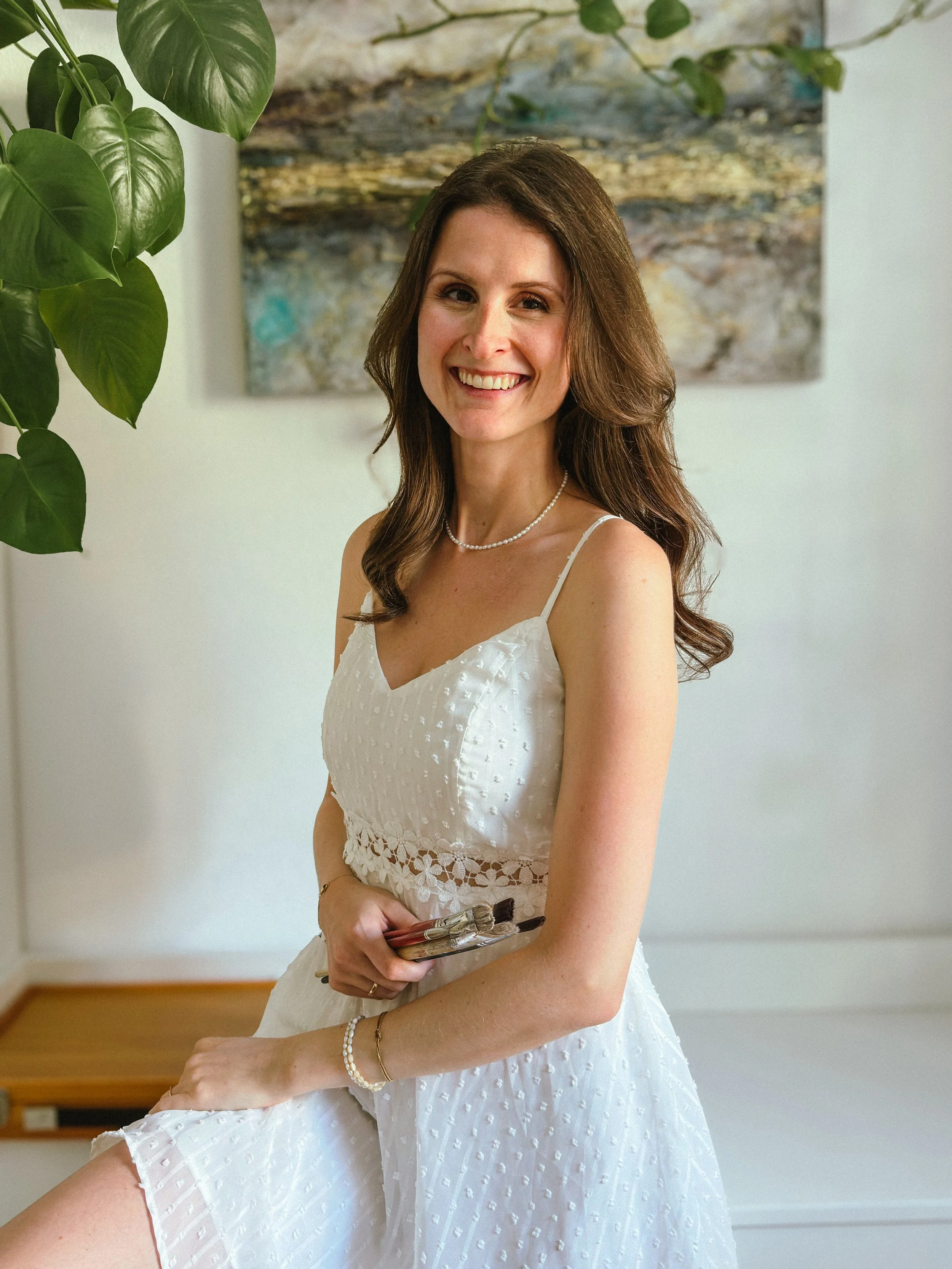 A woman in a white dress with spaghetti straps and lace details, holding makeup brushes, smiling in an indoor setting with a green plant and an abstract painting in the background.