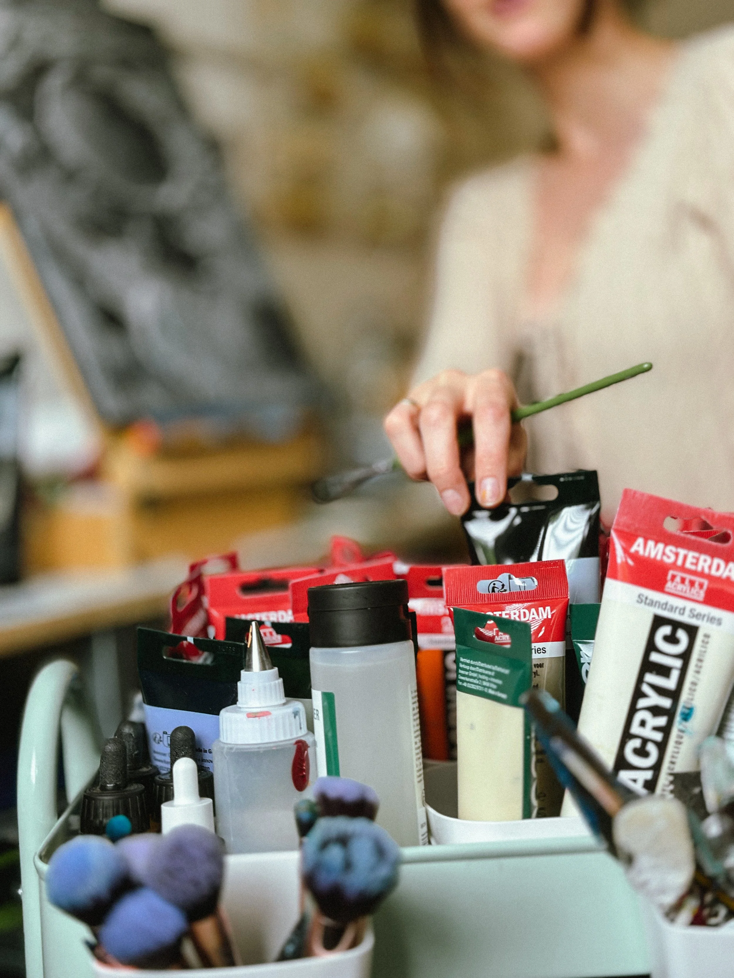 A person is reaching into a drawer filled with art supplies, including paint bottles, brushes, and modeling clay, with drawing and painting tools visible on the table.