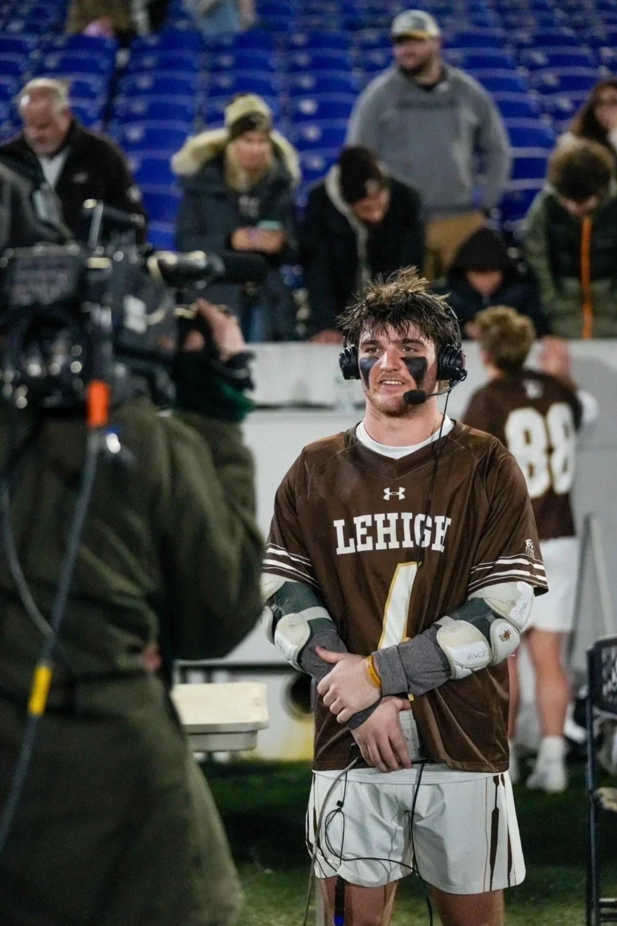 A young male player, wearing a brown Lehigh jersey with the number 1, stands with his arms crossed, talking to a camera crew. He has football gloves and eye black on his face, and he's wearing a headset. In the background, there are spectators and other players, some wearing brown jerseys, in a stadium.
