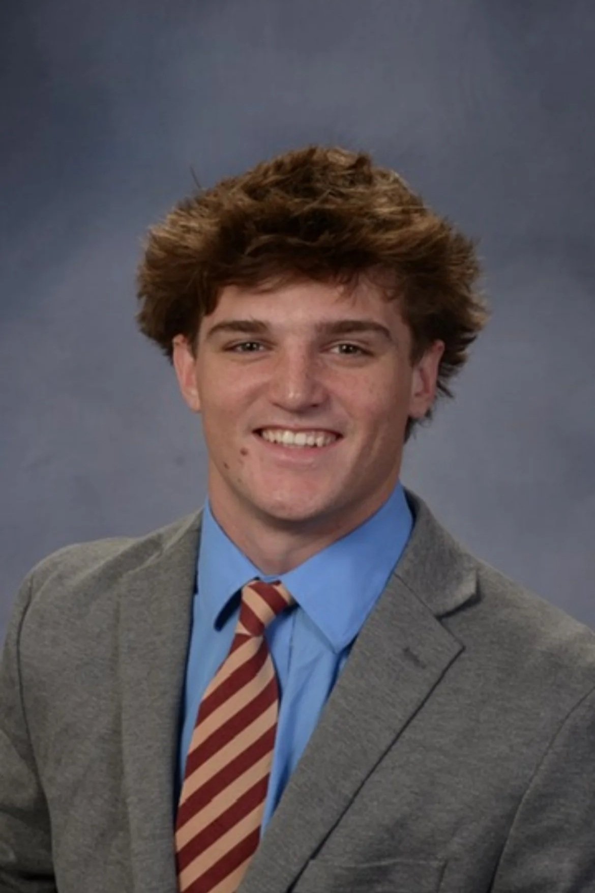 A young man in a gray suit, blue shirt, and striped tie smiling in a professional headshot against a gray background.