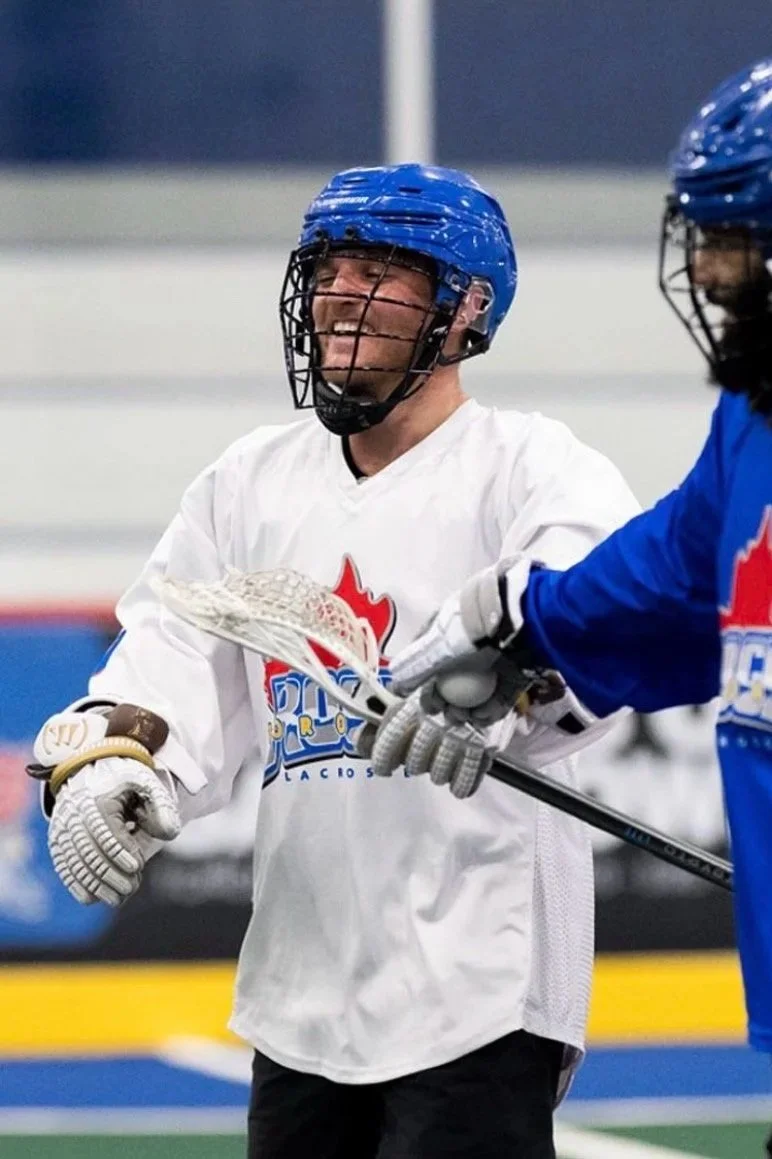 Two men in hockey gear, including helmets, gloves, and jerseys, are on an ice rink. One man, in a white jersey, is smiling and holding a lacrosse stick, while the other, in a blue jersey, is reaching out toward him.