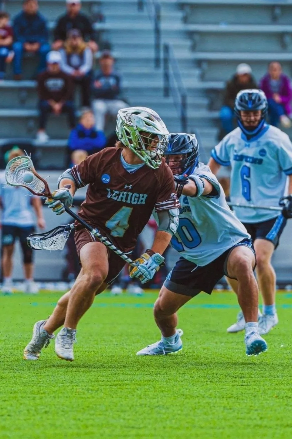 Lacrosse players competing on a grassy field, with one player in a brown jersey and a helmet, and the other in a white jersey and helmet, during a game.