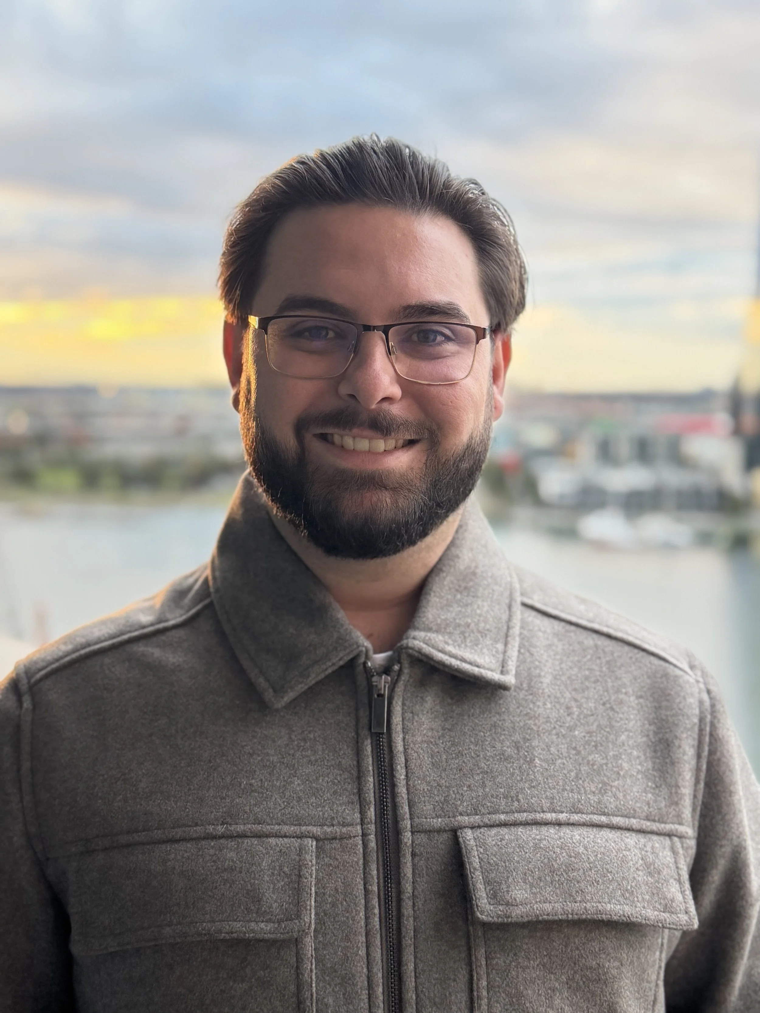 A man with glasses and a beard smiling in front of a background of the city skyline and water, wearing a gray zip-up jacket.
