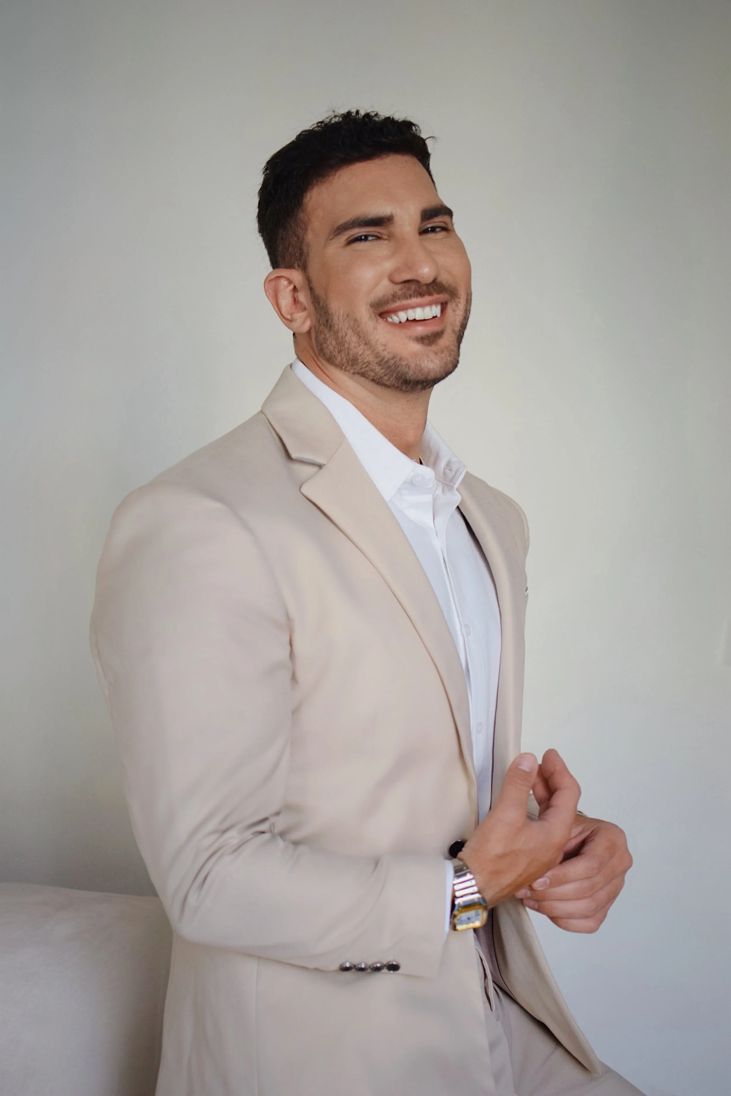 Hombre de piel clara, cabello oscuro y barba, vestido con saco beige y camisa blanca, sonriendo y mostrando una actitud confiada, con fondo liso y neutro.