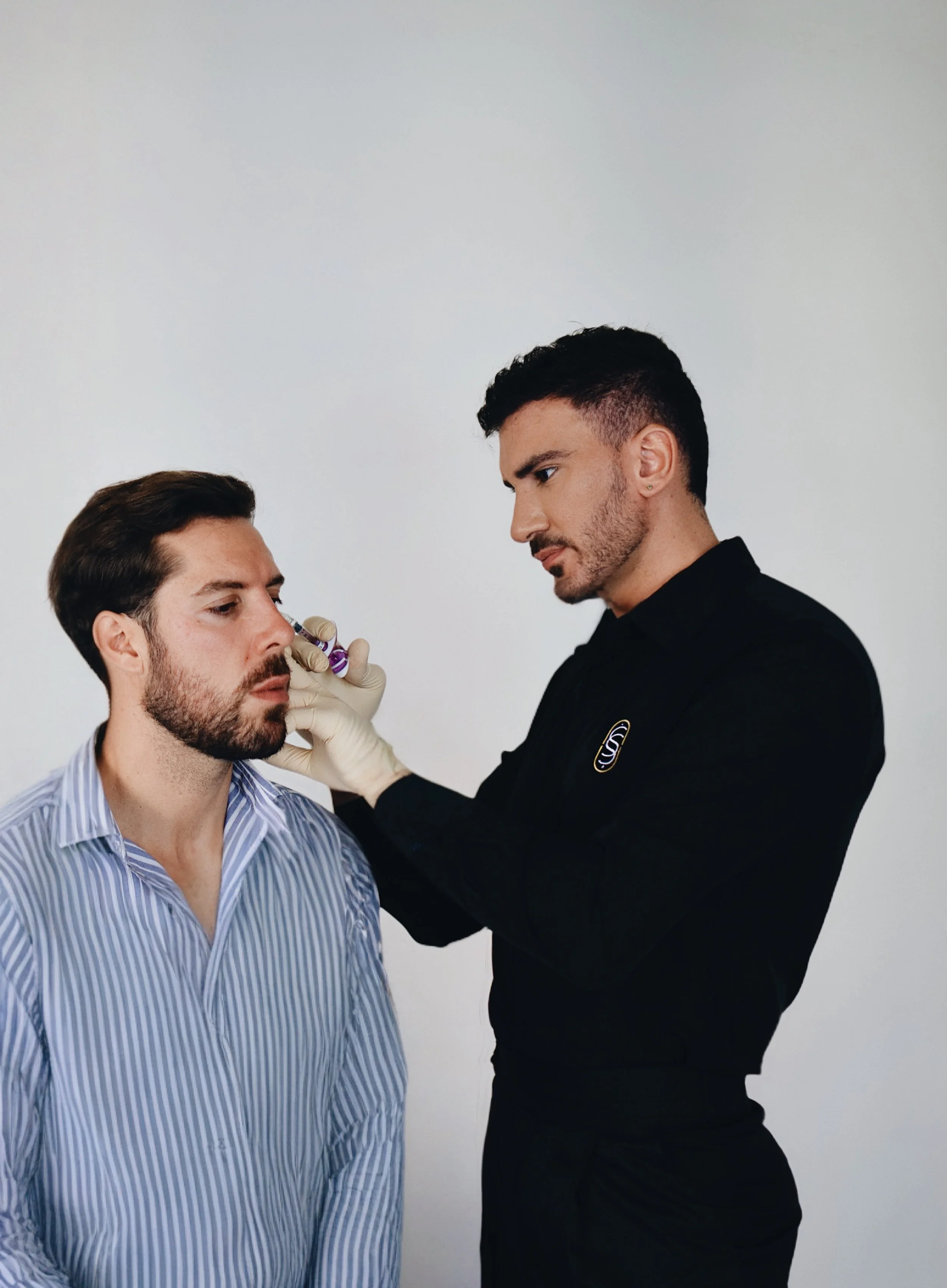 Un hombre con barba y cabello oscuro, vestido con camisa a rayas, recibe una vacuna de una profesional de la salud con guantes. La profesional tiene el cabello oscuro y usa un uniforme negro. La escena ocurre en un fondo blanco.