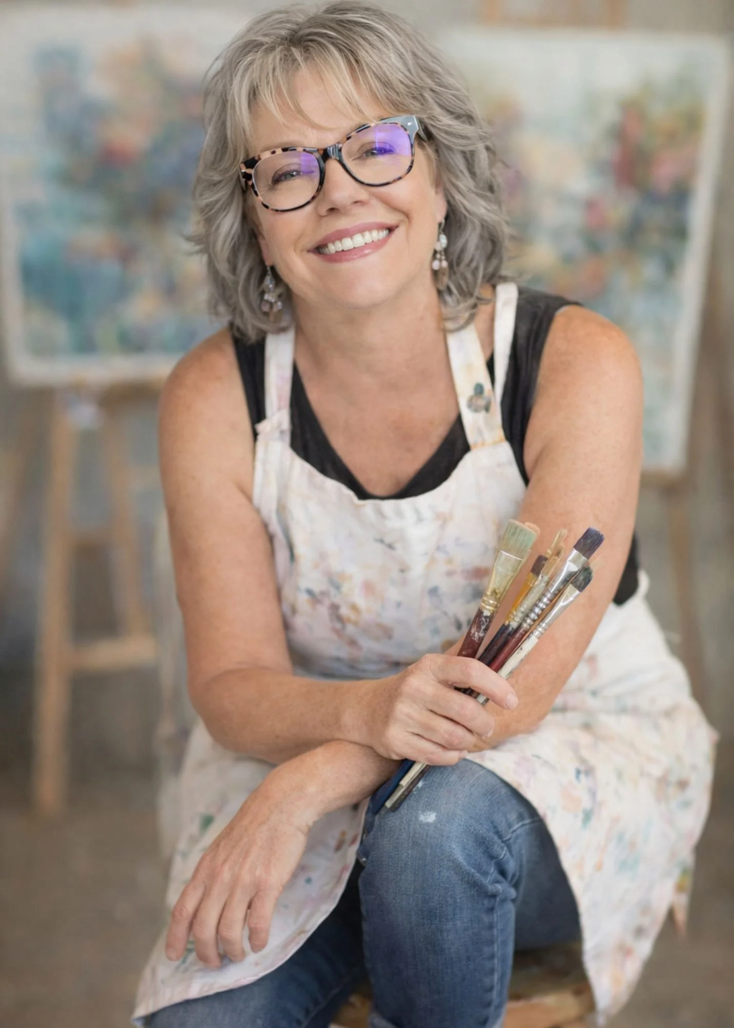 Picture of Kimbella Buckman sitting on a stool with paint apron and paint brushes smiling.