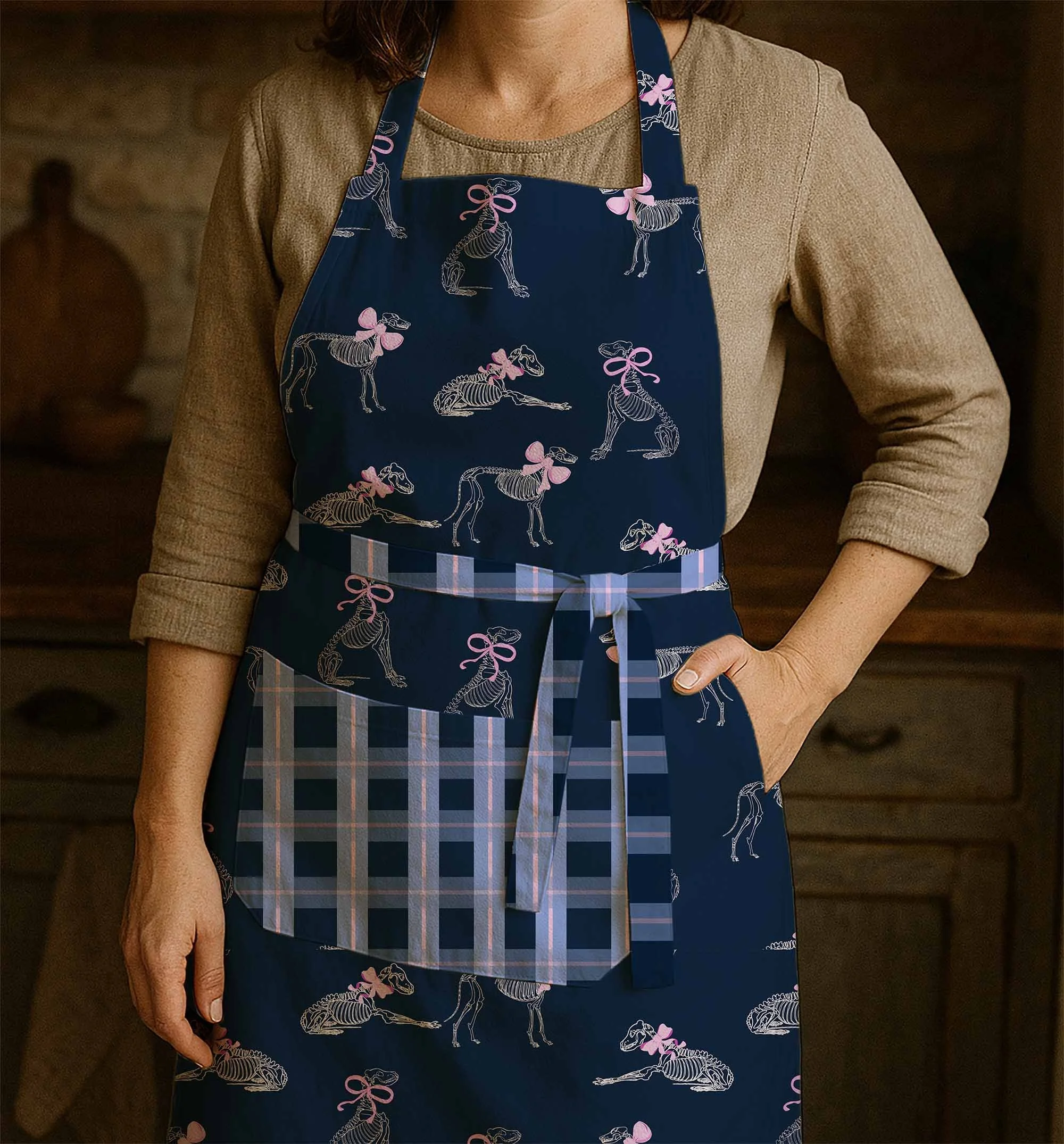 A person wearing a tan long-sleeve shirt with an apron that has a skeleton and bow design on it, standing indoors.