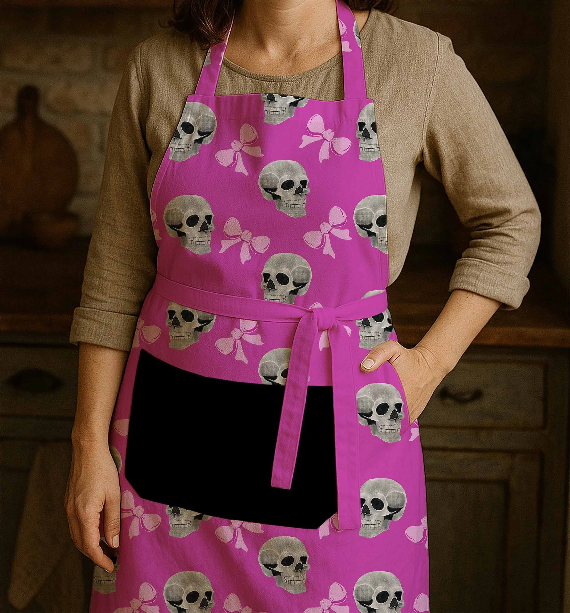 Woman wearing a beige shirt with a pink apron decorated with skulls and bows, standing in front of a dark background.