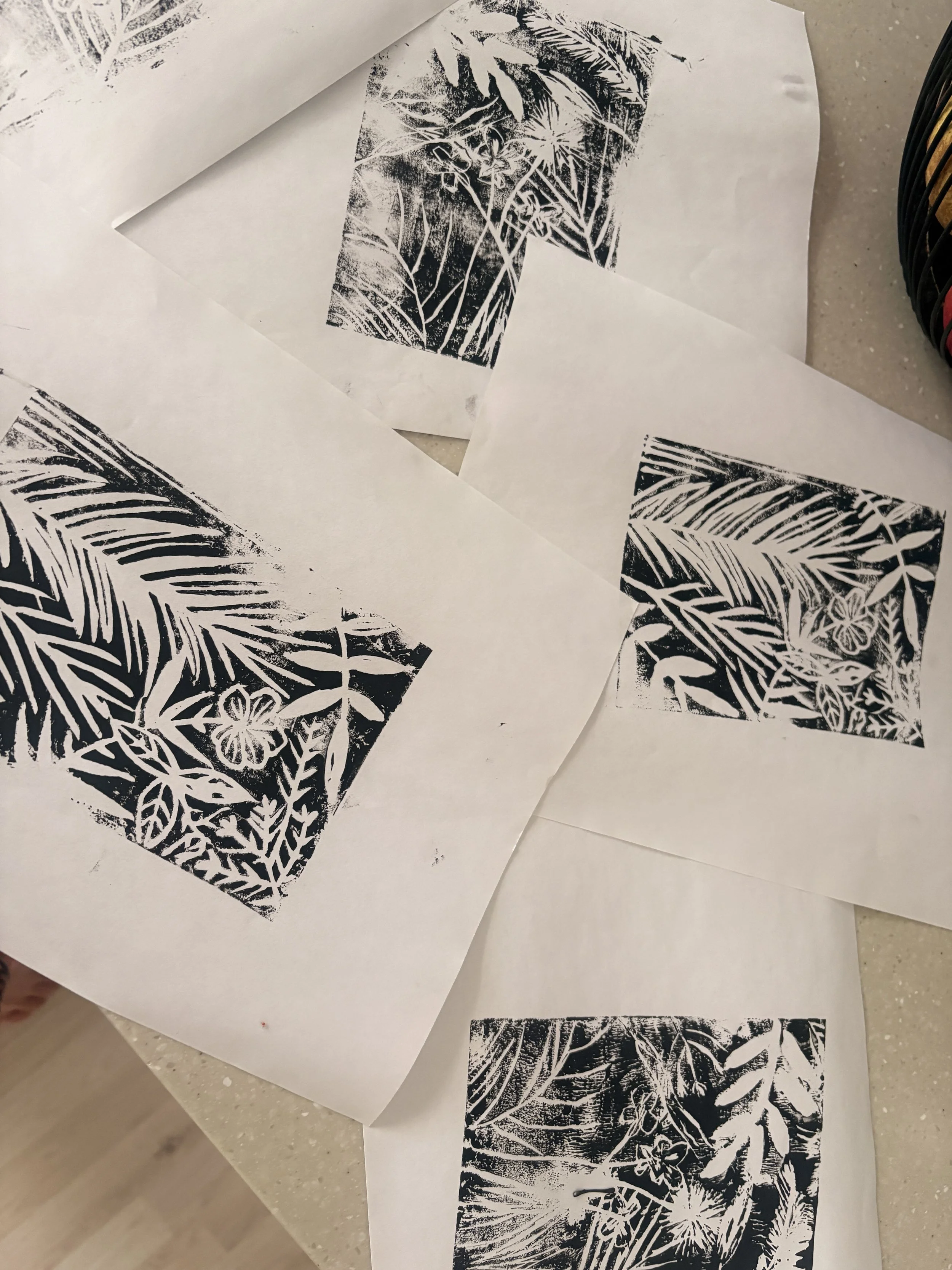 Black and white botanical printmaking sketches on white paper, showing various plant leaves and floral designs, some stacked or overlapping on a light-colored surface with a black wire basket visible on the side.