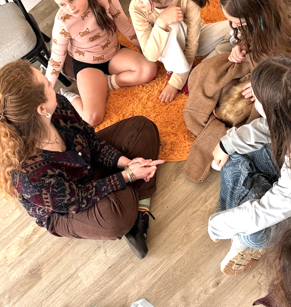 Group of children and an adult sitting on the floor in a circle, engaging in conversation.