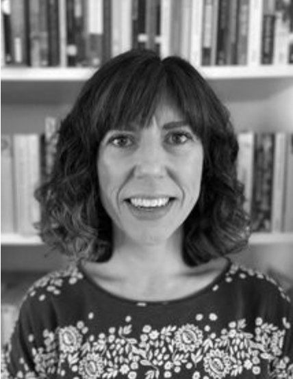 Black and white portrait of a woman with shoulder-length wavy hair, smiling, in front of shelves filled with books.