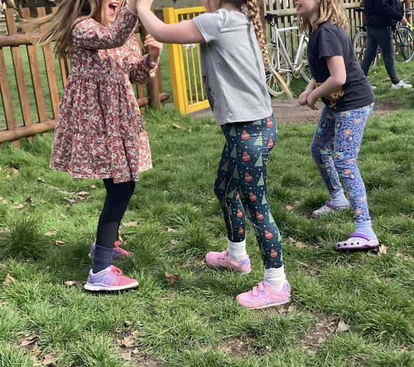 Three young girls playing and having fun outdoors on green grass, with a wooden fence, bicycle, and a person in the background. One girl is wearing a floral dress and pink sneakers, while the other two are in patterned leggings and sneakers.