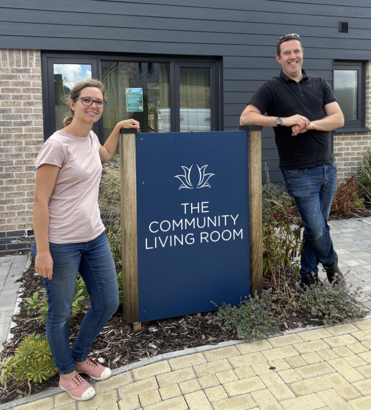 Two people standing next to a sign for The Community Living Room in front of a modern building with brick and gray siding.