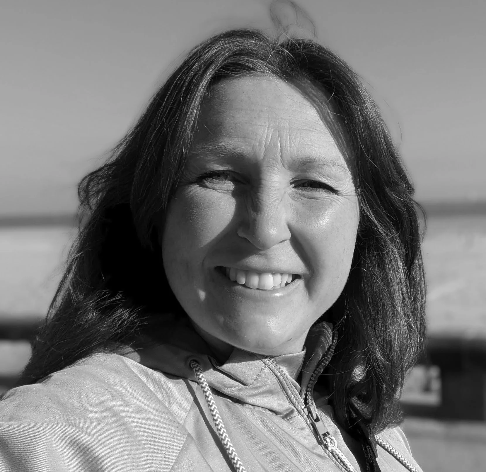 A black and white close-up of a smiling woman outdoors in a zip-up jacket with a rope-like detail, with a blurred background and open sky.