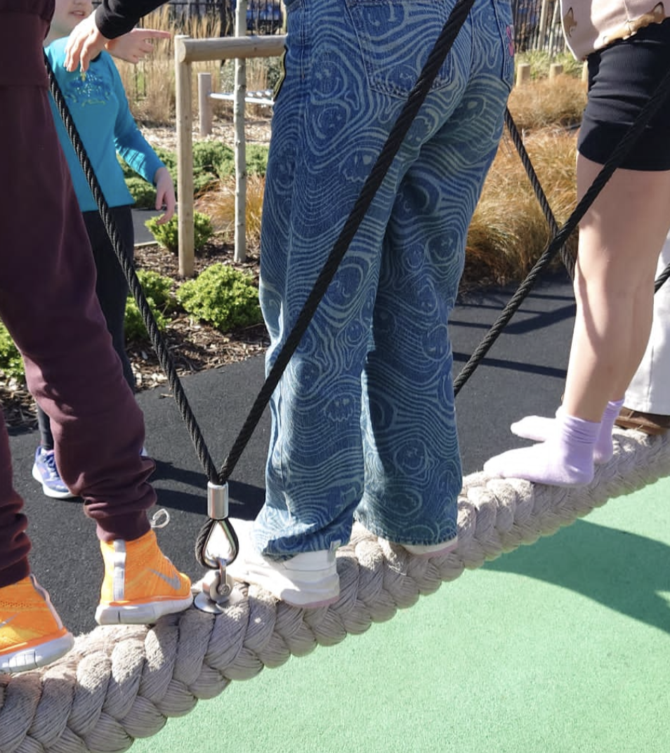 Close-up of children walking on a thick rope bridge outdoors, with shoes and colorful clothing visible.