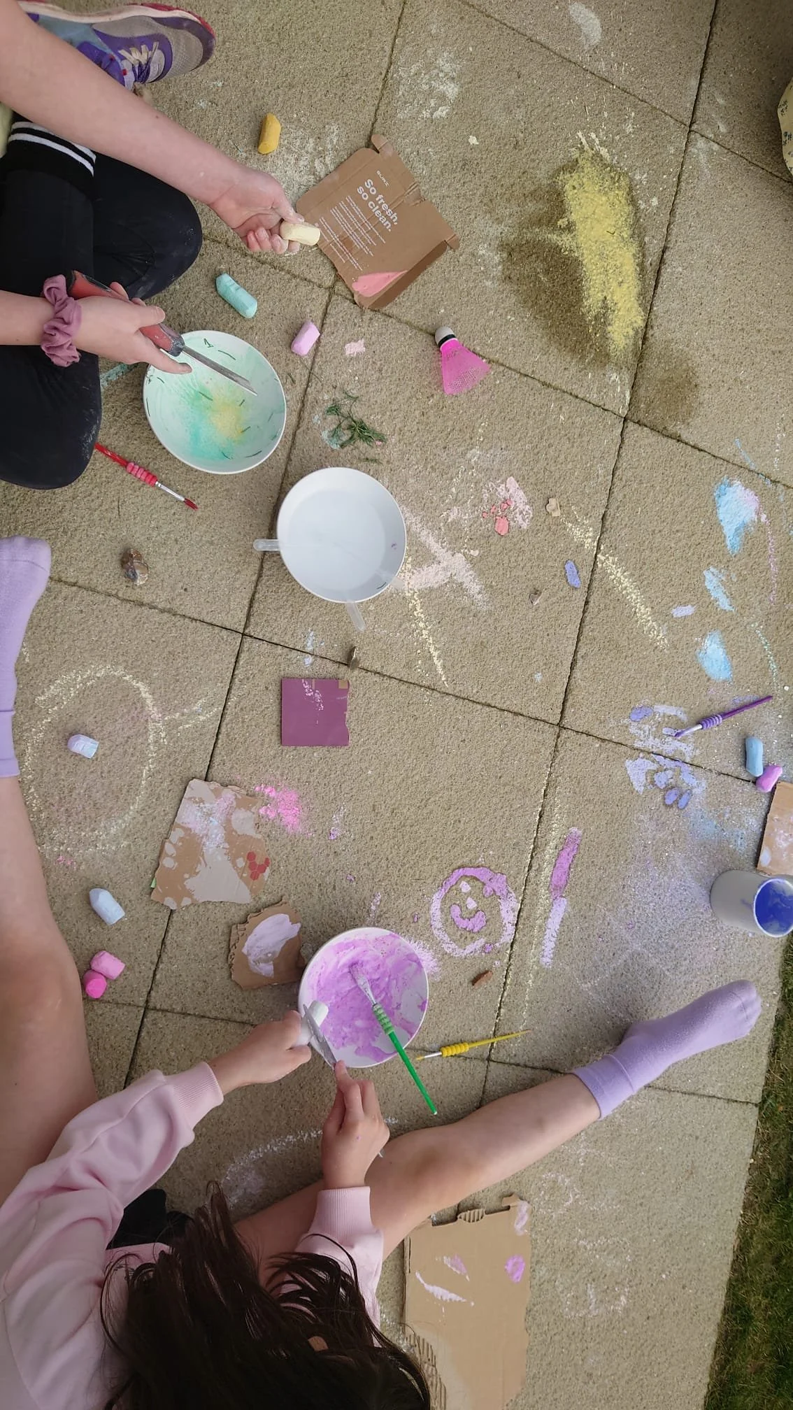 Children playing with chalk and chalk dust on a concrete patio, with some cups and art supplies scattered around.