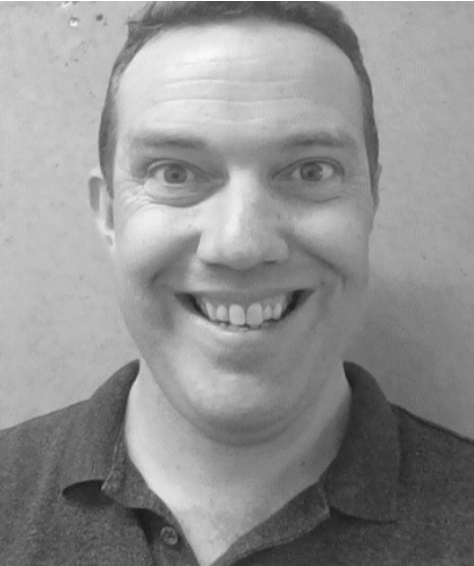 Black and white close-up photo of a smiling man with short hair, wearing a collared shirt, standing against a plain wall.