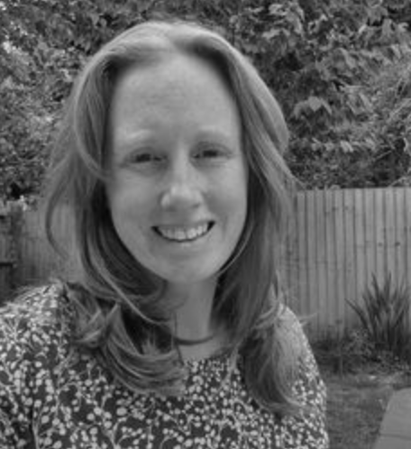 A smiling woman with shoulder-length hair outdoors, in front of a wooden fence and trees.