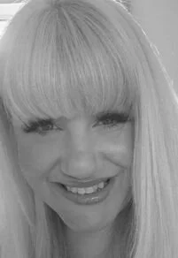 Close-up of a woman with long blonde hair and bangs, smiling at the camera in black and white.