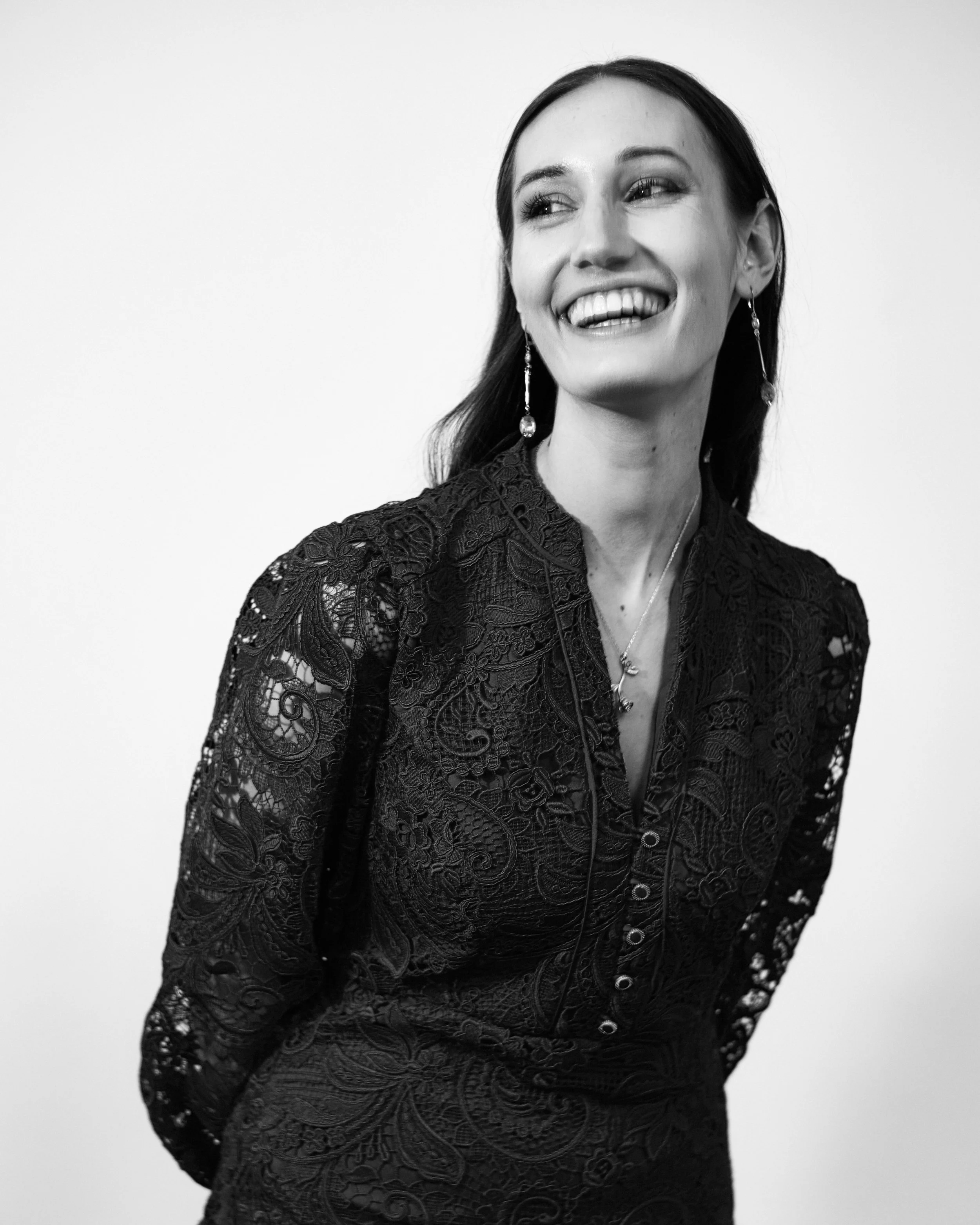 Black and white photo of a woman with long dark hair, wearing a lace dress and dangling earrings, smiling and looking slightly to the right.