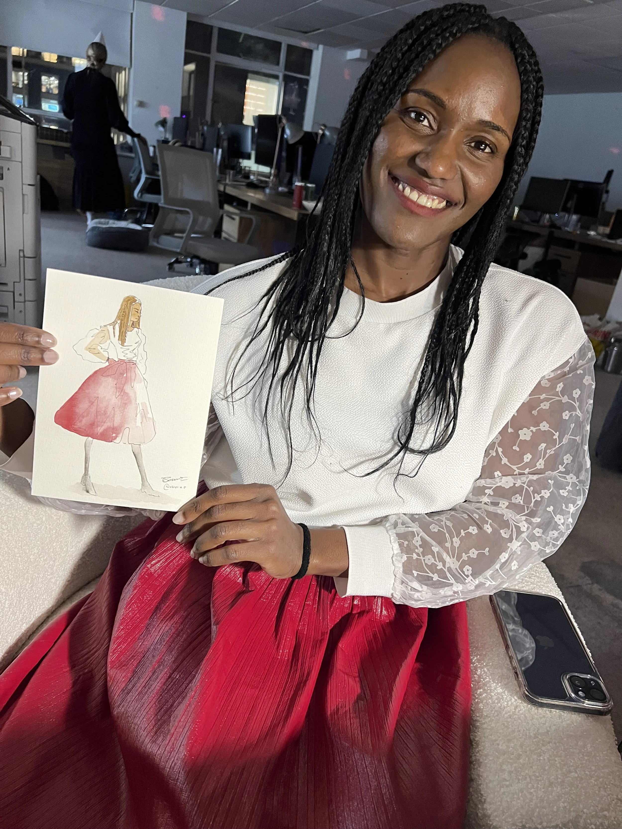 A smiling woman with braided hair holding a fashion illustration of a woman in a white top and red skirt. The woman is sitting on a couch in an office or studio space with computers and office chairs in the background.