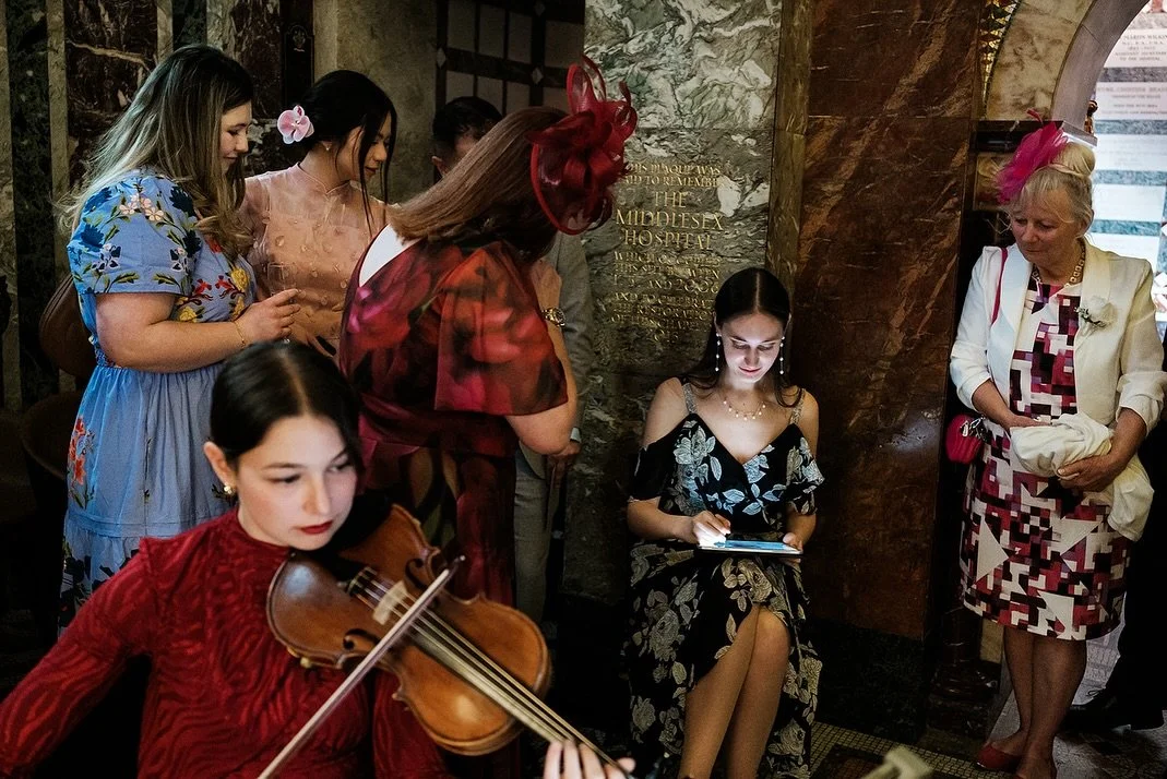 A group of women, some dressed in floral dresses, gather around a young woman playing the violin, while another woman with a flower in her hair looks at her tablet, in an elegant indoor setting.