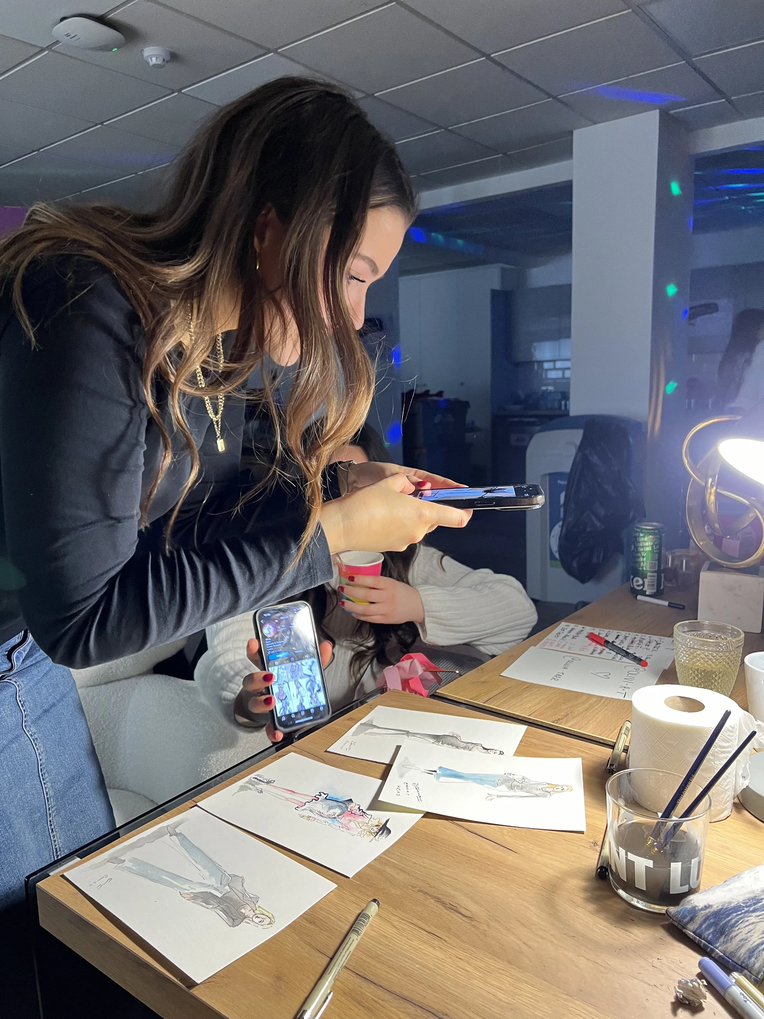 A woman with long brown hair, wearing a black jacket and gold jewelry, is looking at her phone while seated at a table with sketches of fashion illustrations. There are pens, papers, and a glass of water on the table, and other people are partially v