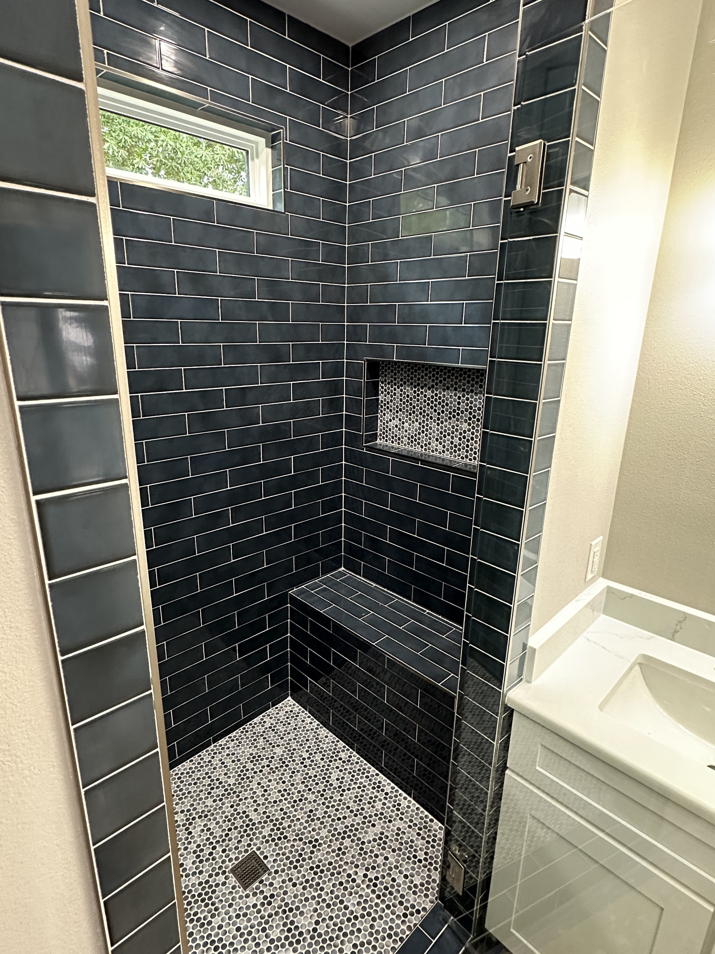 A modern shower area with black subway tile walls, a small rectangular window, black and white mosaic tile floor, and a built-in bench with mosaic tile surface. There's a recessed shelf with white mosaic tiles, and a strip of glass blocks on the left side.