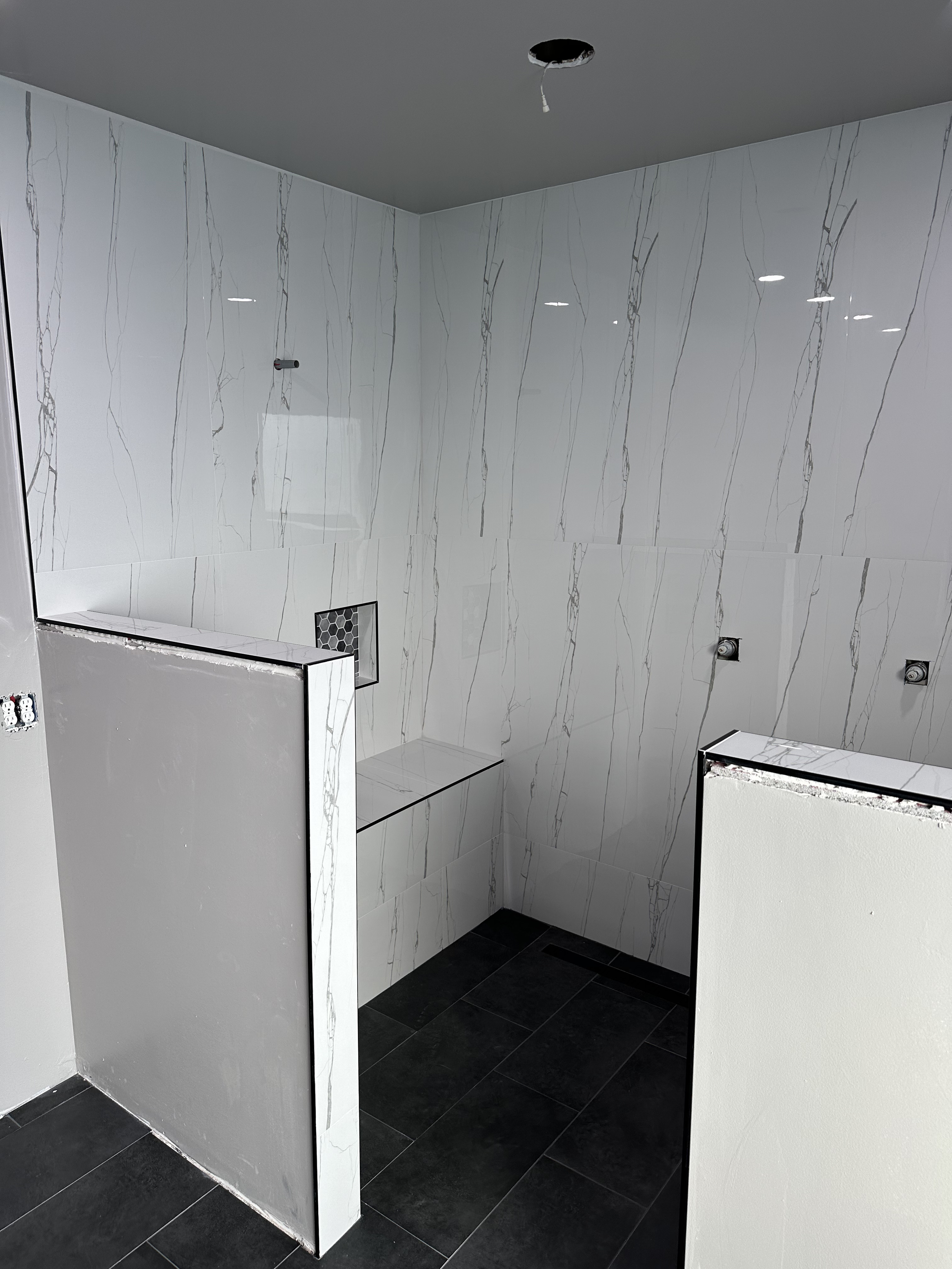 Interior of a bathroom or shower area with white marble tiles with gray veining, black flooring, and uninstalled fixtures, including an opening in the ceiling for a light or vent and electrical outlets on the walls.